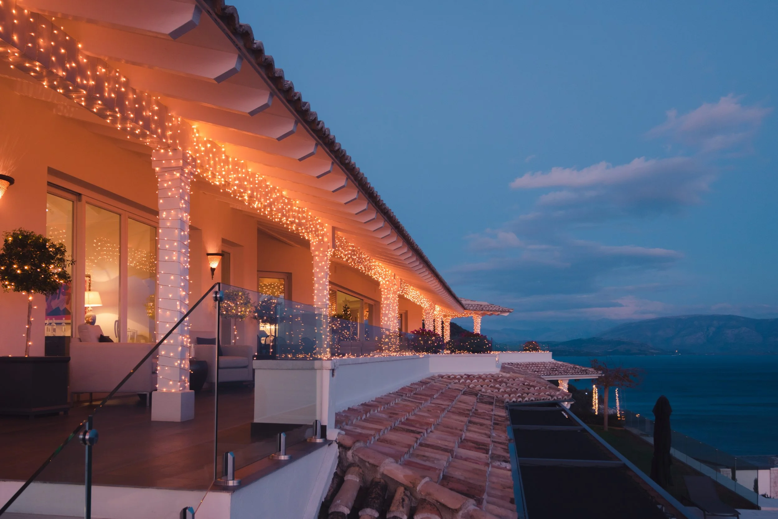 A waterfront house decorated with string lights on the balcony during dusk, overlooking the water and distant mountains.