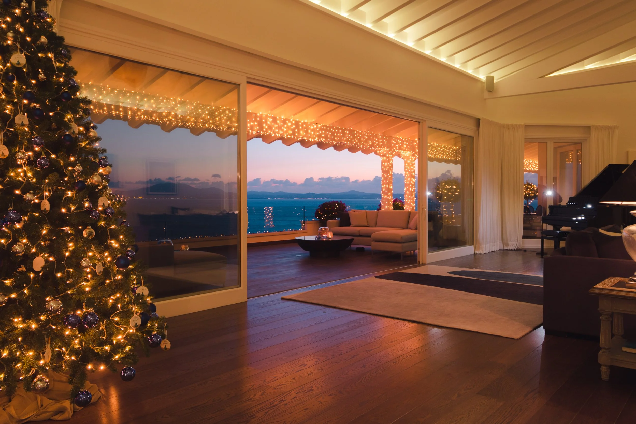 A living room decorated for Christmas with a large Christmas tree, fairy lights, and a cozy seating area on a balcony overlooking a body of water at sunset.