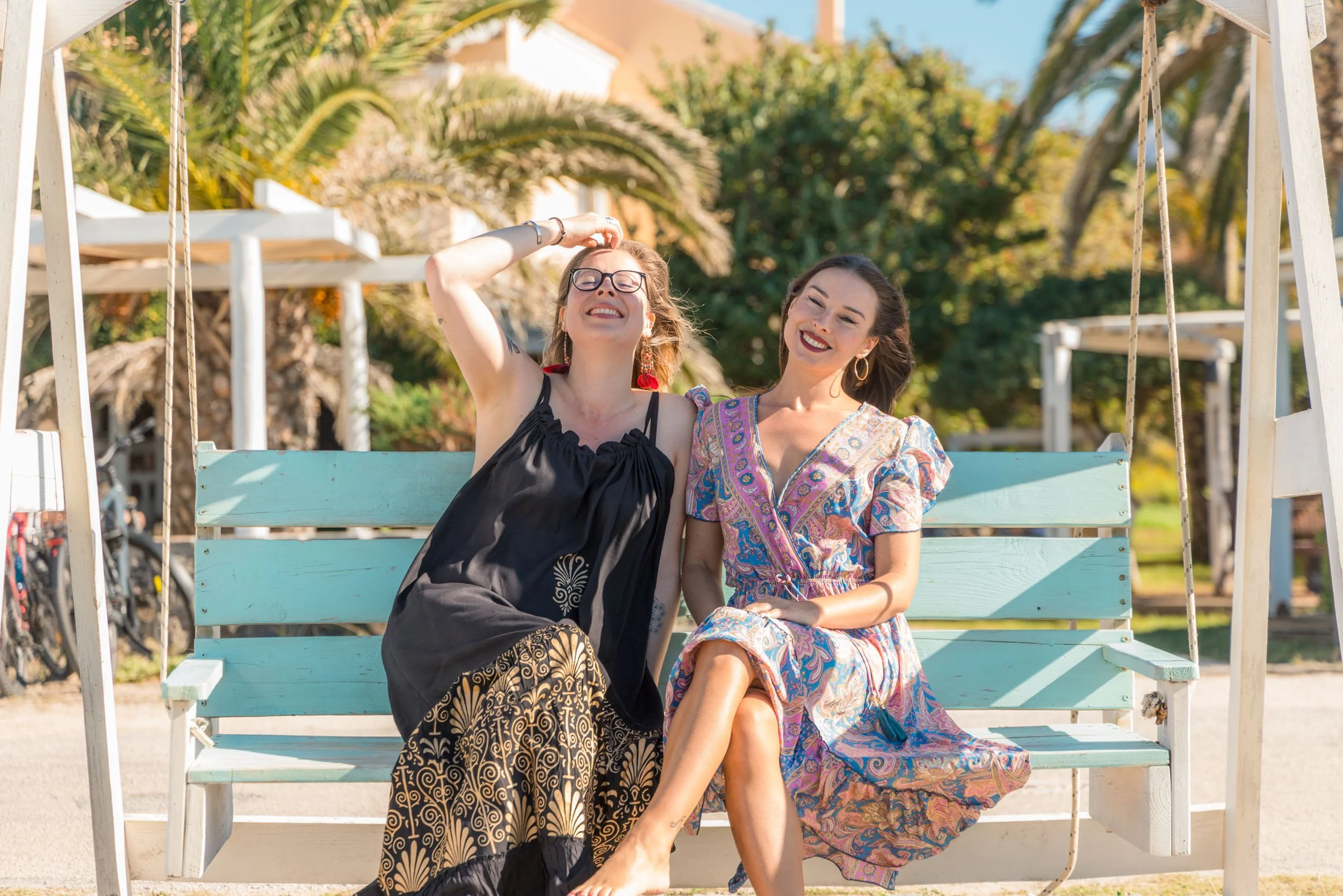 Two women sitting on a swing, smiling and enjoying a sunny day outdoors with trees and palm plants in the background.