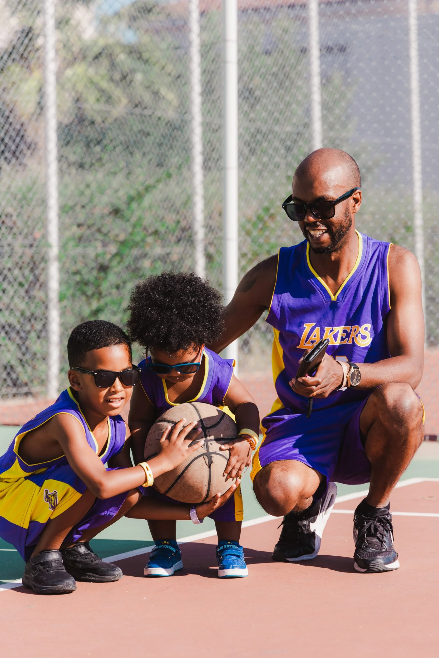 A basketball coach crouching on an outdoor basketball court with three young children, two wearing sunglasses and one holding a basketball, all wearing Los Angeles Lakers jerseys.