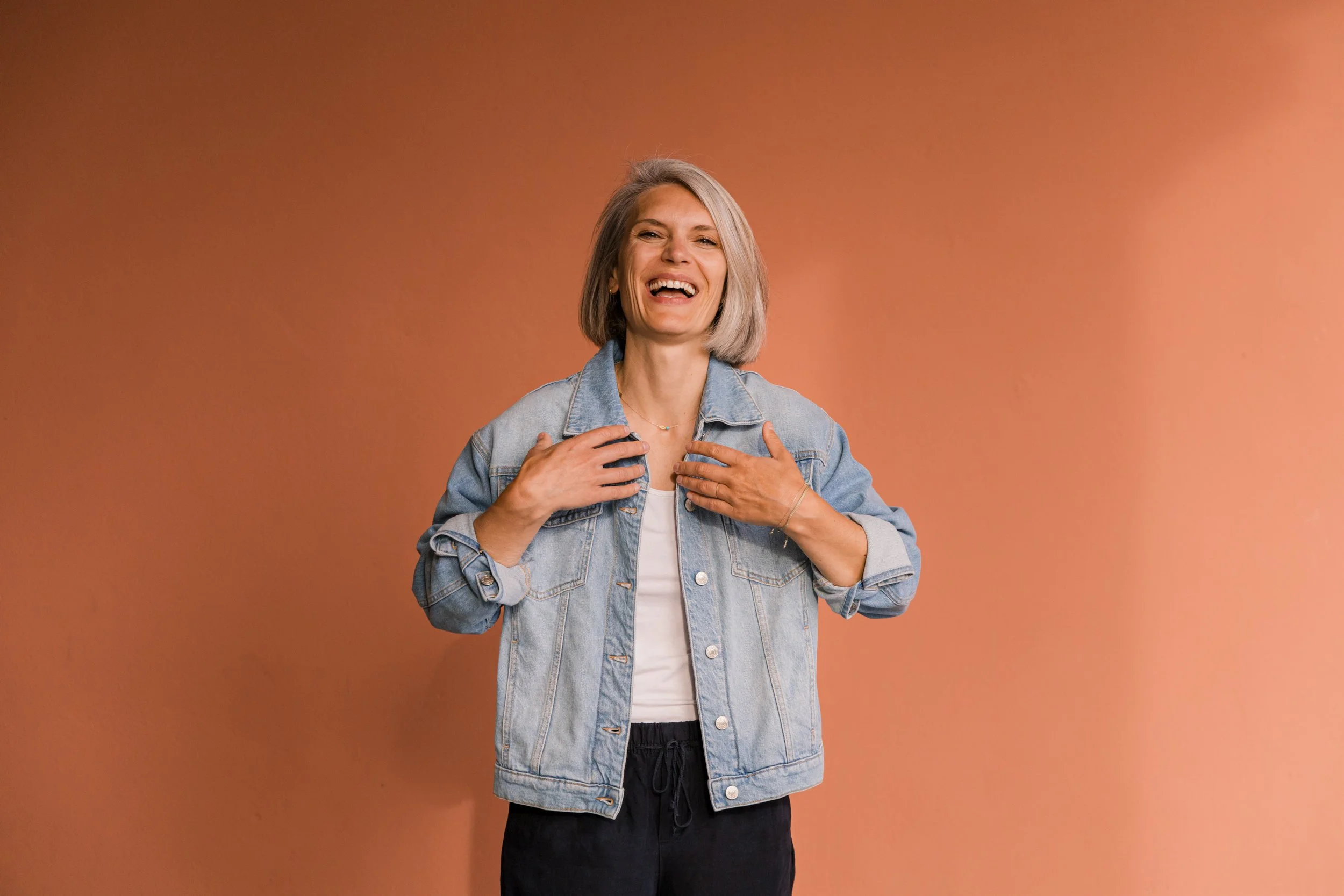 A smiling woman with gray hair wearing a denim jacket and white shirt against a peach-colored background.