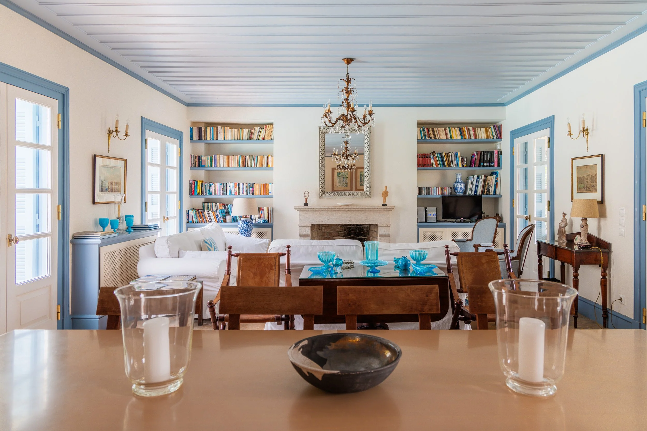 A bright living room with light-colored walls, blue trim, a chandelier, a fireplace, and built-in bookshelves filled with books. There is a large white sofa, a glass dining table with blue glassware, and various decorative items.