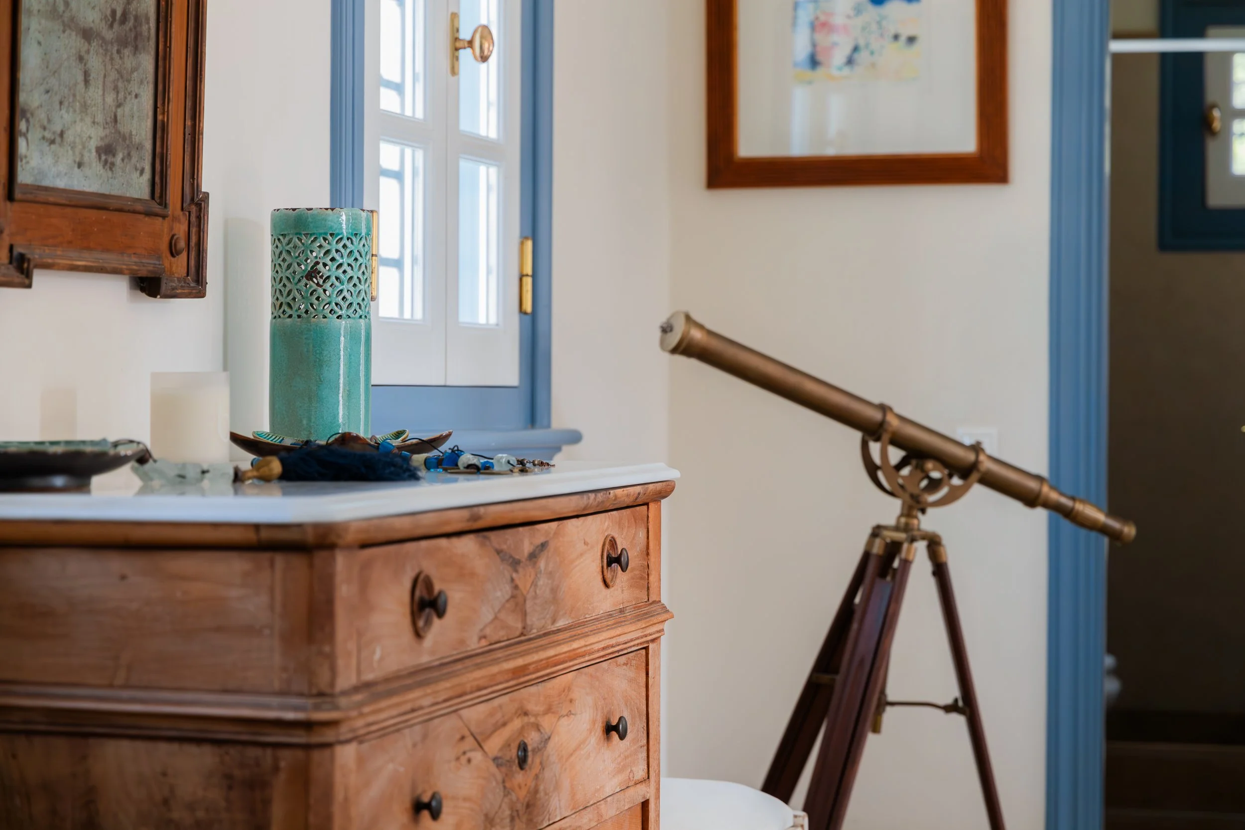 A room corner with a vintage wooden dresser, a green decorative vase, a small sculpture, and jewelry on top. There is a large telescope on a wooden tripod pointed towards the window. The window has a blue frame, and the wall has framed artwork.