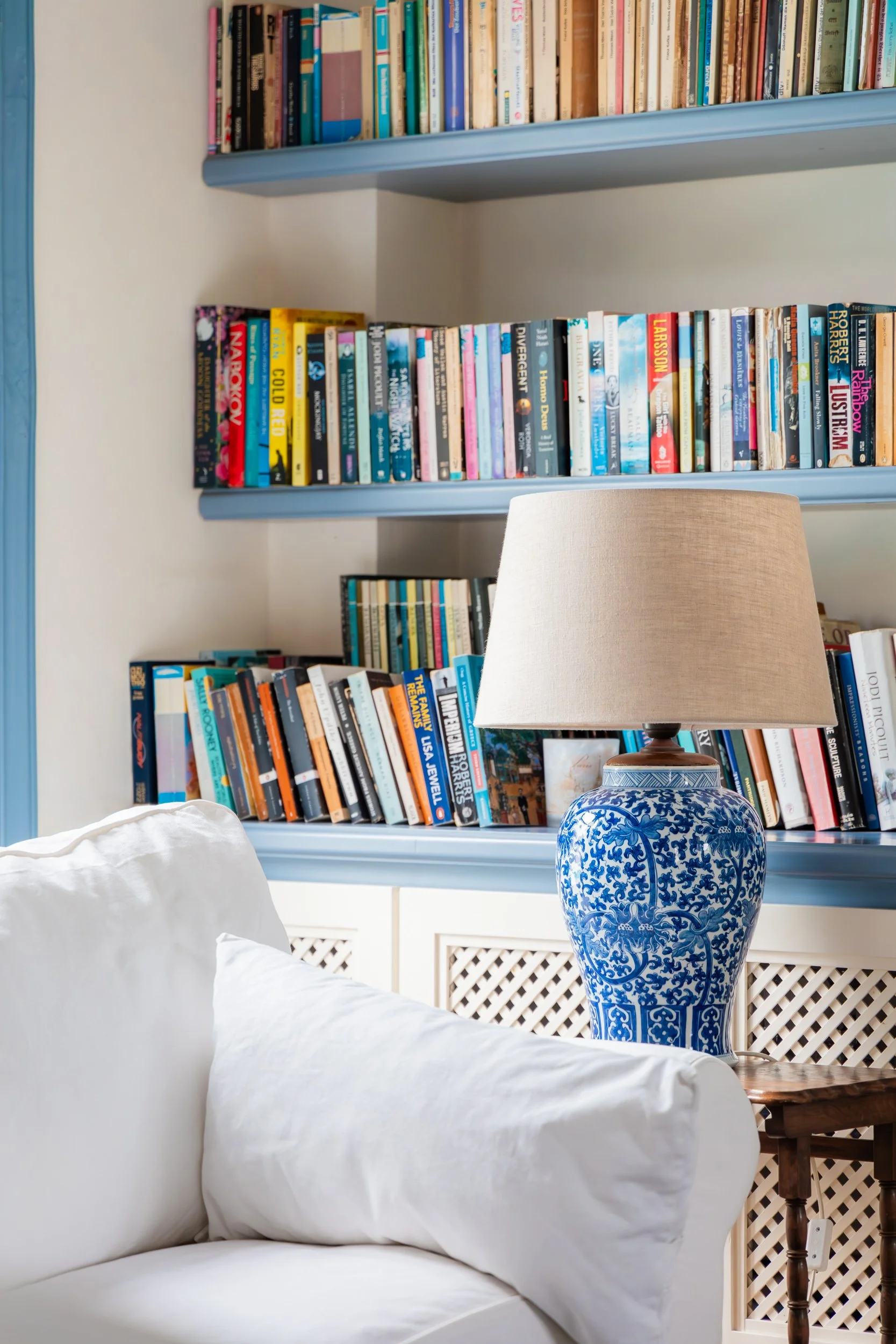 A cozy living room corner with a white couch, a blue and white ceramic lamp with a beige shade, and two blue bookshelves filled with various books.