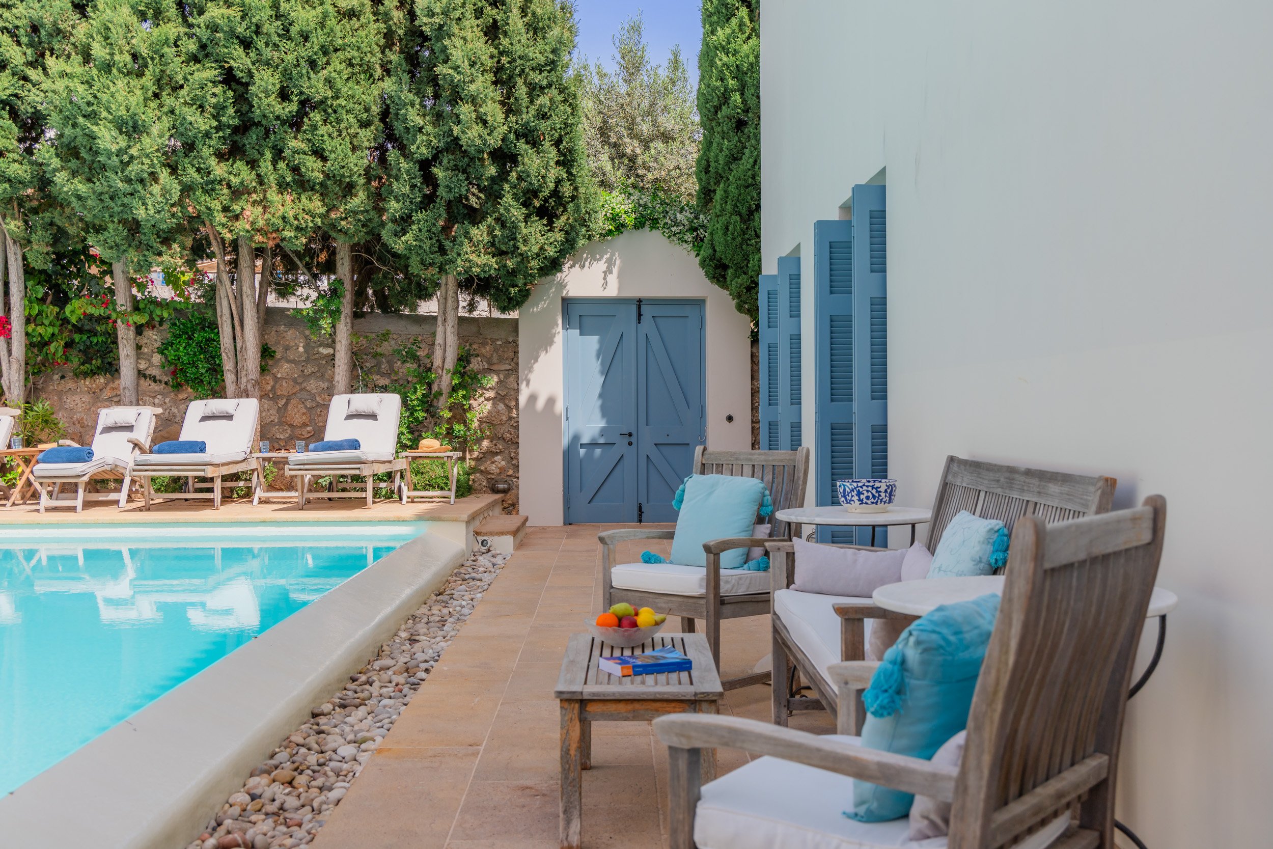 Outdoor poolside seating area with wooden chairs, cushions, a small table with a bowl of fruit, and lounge chairs in the background with towels, surrounded by lush green trees and a stone wall.