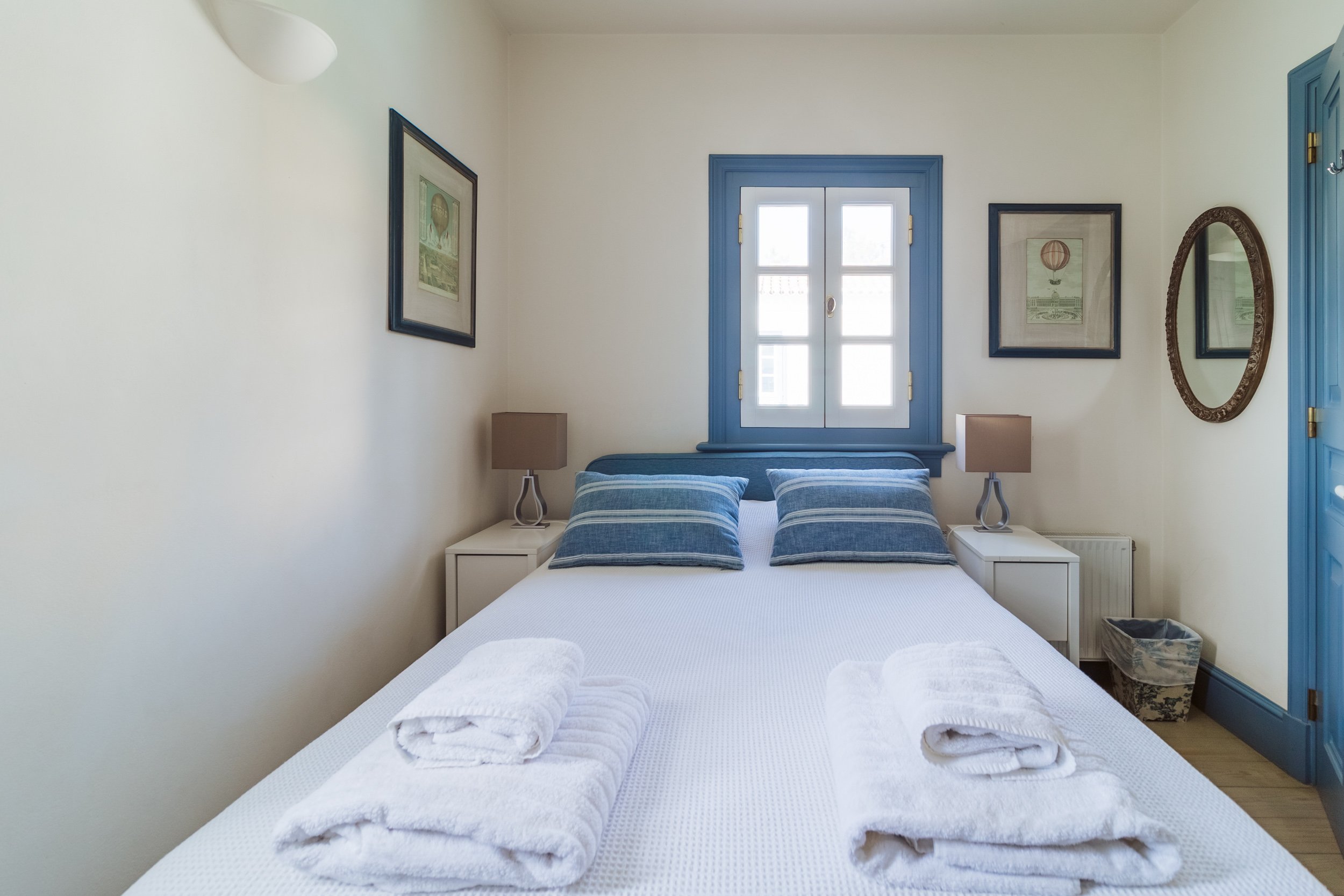 A cozy bedroom with a white bed, blue-striped pillows, and blue window trim. Two white nightstands with beige lamps flank the bed. The room has light-colored walls, framed artwork, and a mirror. Towels are neatly folded on the bed.