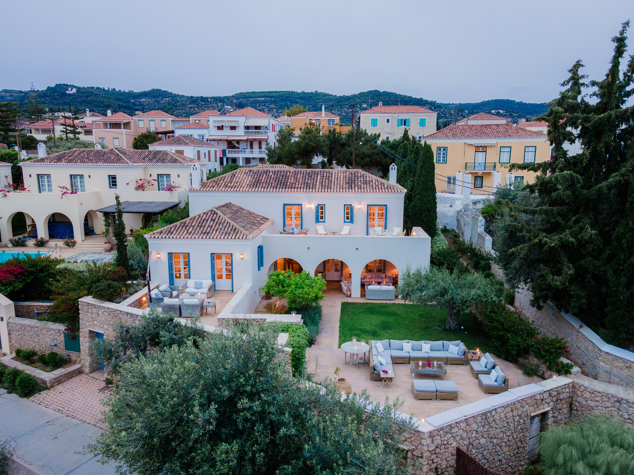Aerial night view of a Mediterranean style house with outdoor seating area, surrounded by greenery and neighboring houses.