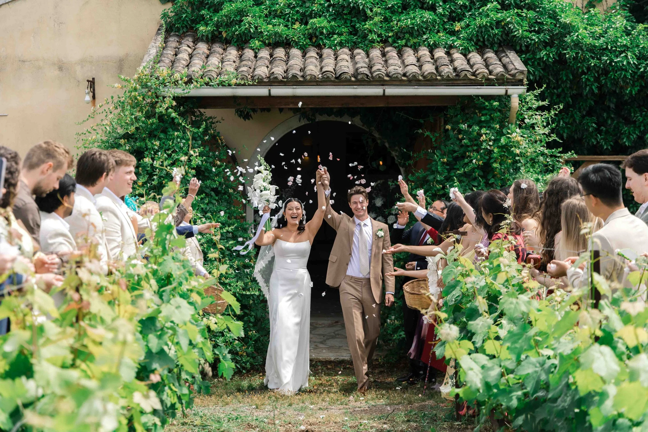 A bride and groom walking through a crowd of wedding guests under an archway, celebrating with confetti.