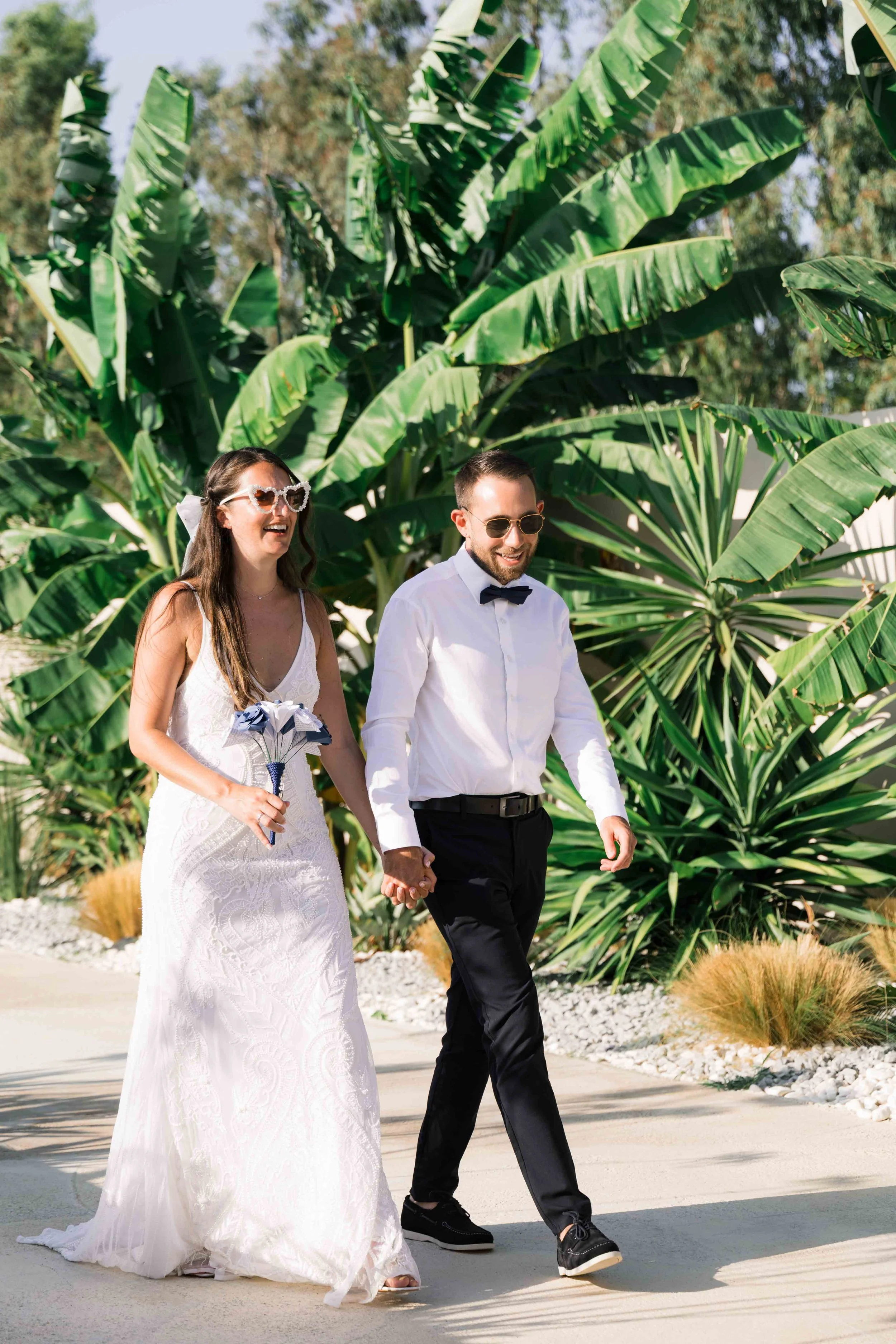 A smiling couple dressed in wedding attire holding hands and walking outdoors in front of lush green plants, with the woman wearing a white wedding dress and sunglasses, and the man wearing a white shirt, black pants, bow tie, and sunglasses.