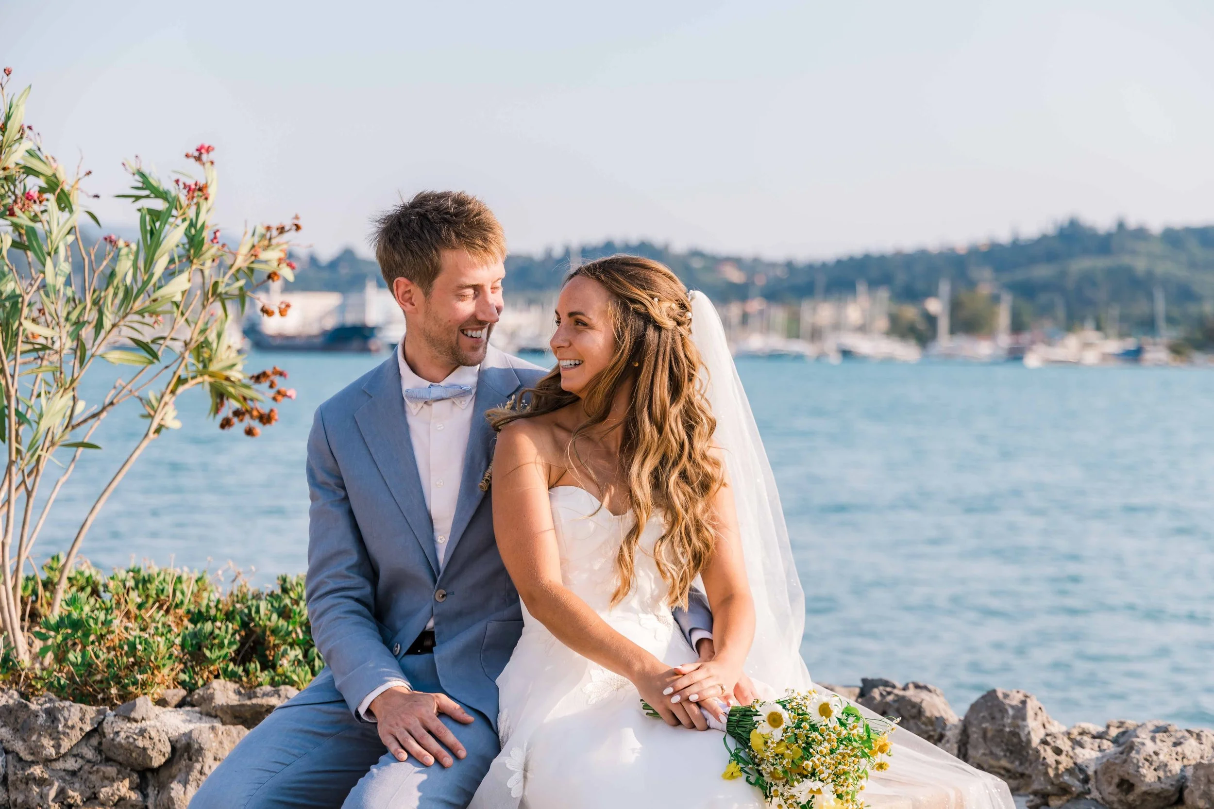 A newlywed couple sitting by the water, smiling and looking at each other, with a bouquet of yellow and white flowers, against a background of boats and hills.