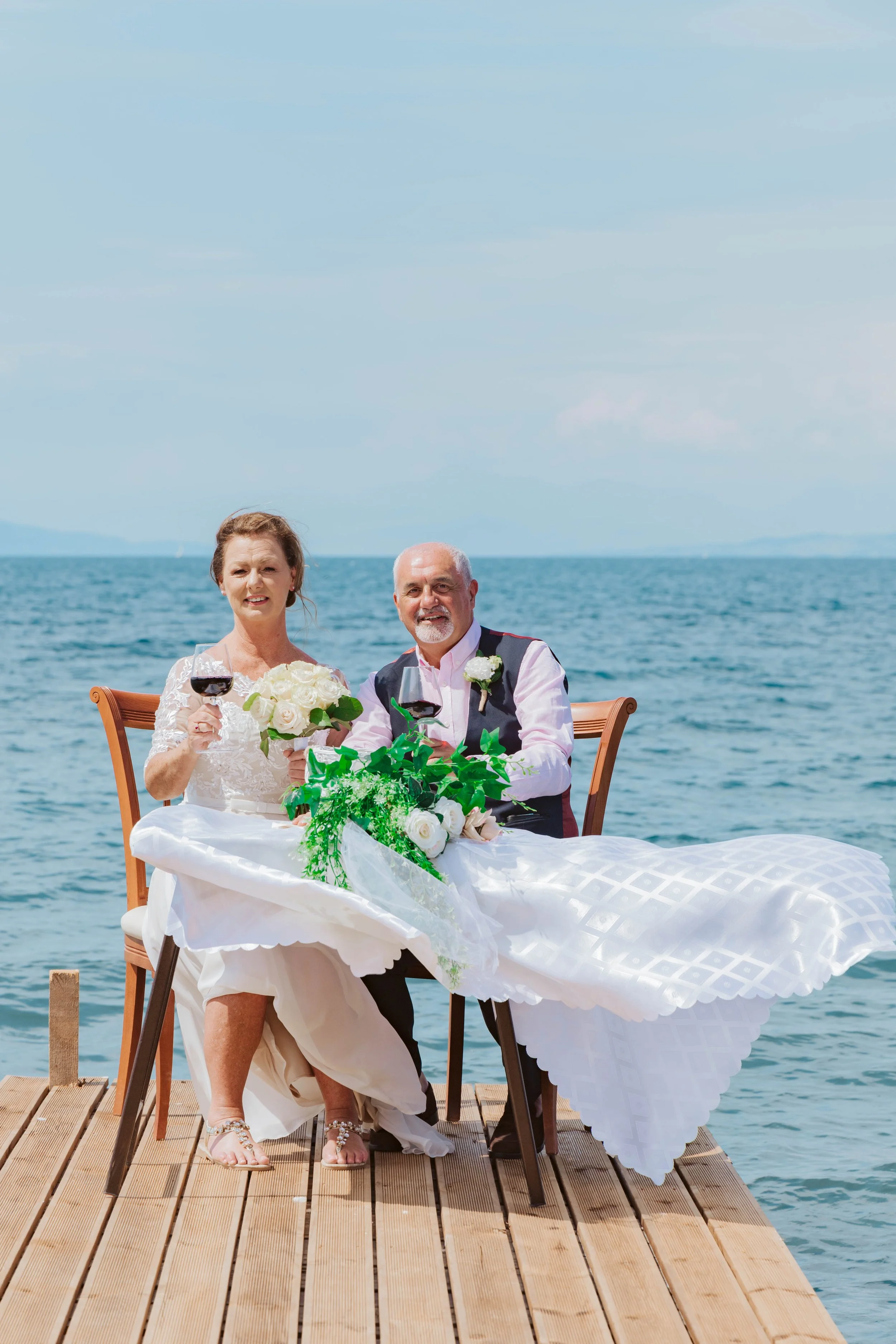 Senior couple dressed in wedding attire sitting at a table on a pier by the water, holding wine glasses and smiling, during a celebration.