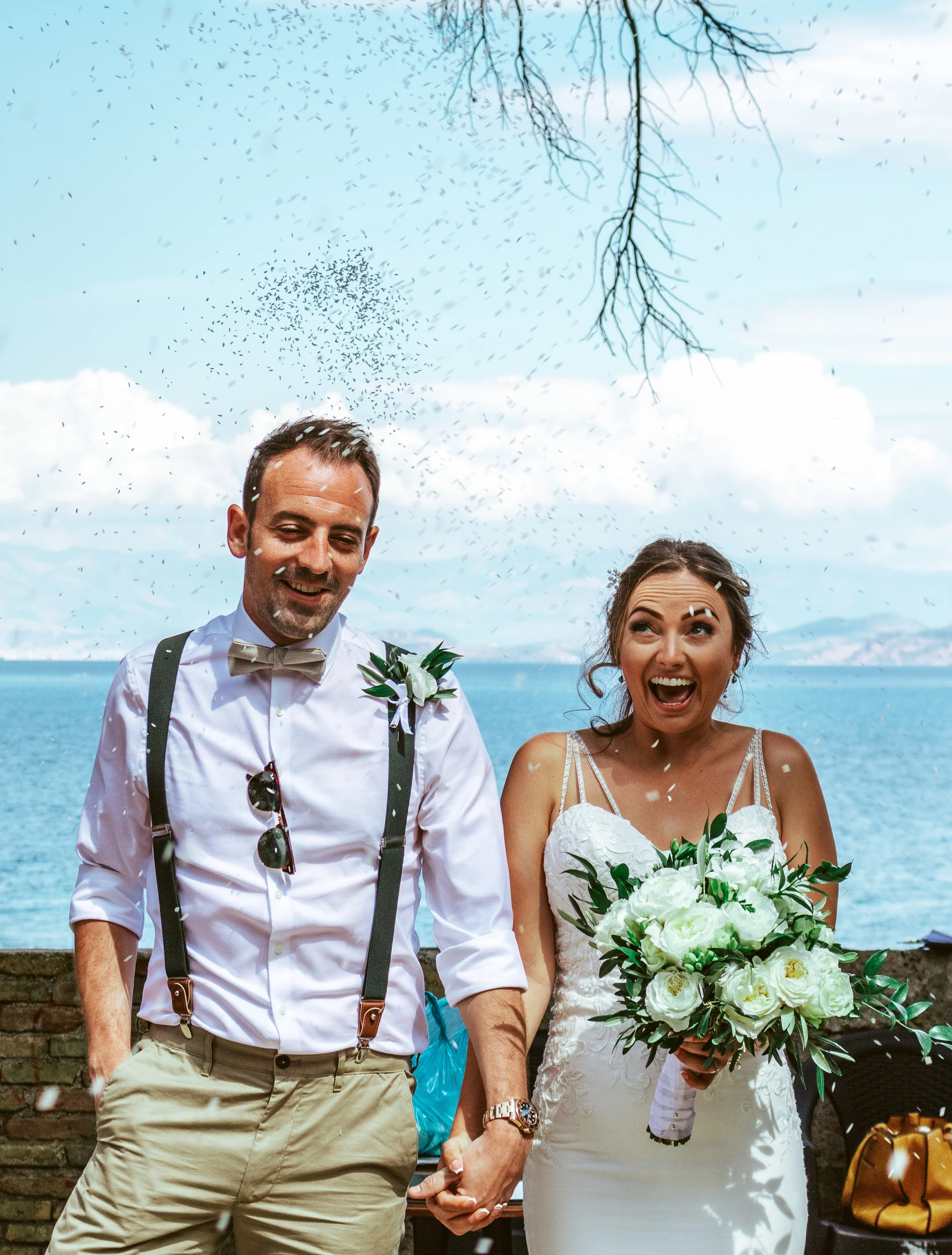 A bride and groom holding hands and smiling at each other outdoors near the water, with confetti falling from above on a bright day.