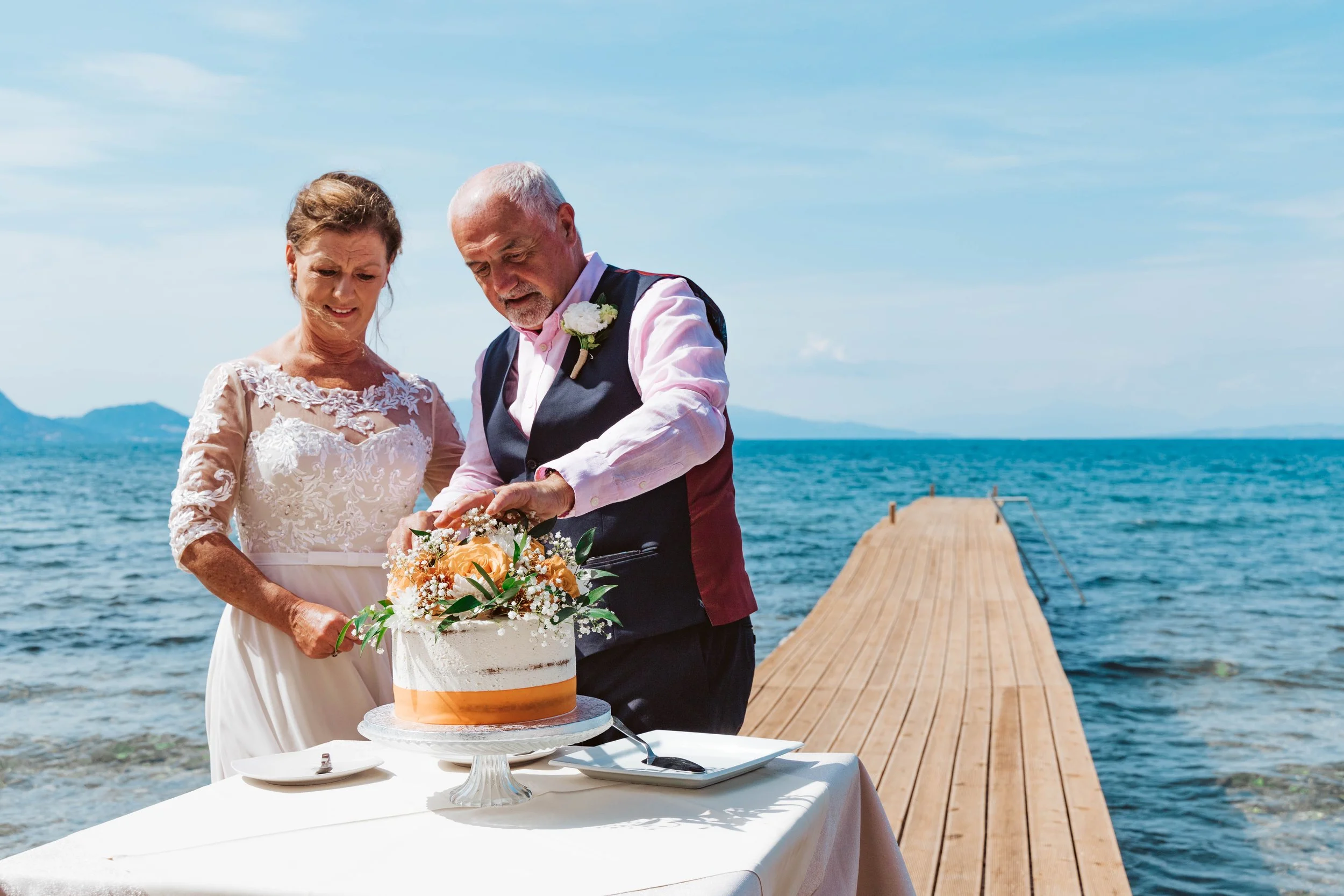 An elderly couple is cutting a wedding cake on a table outside by the ocean, with a wooden pier extending into the water in the background.