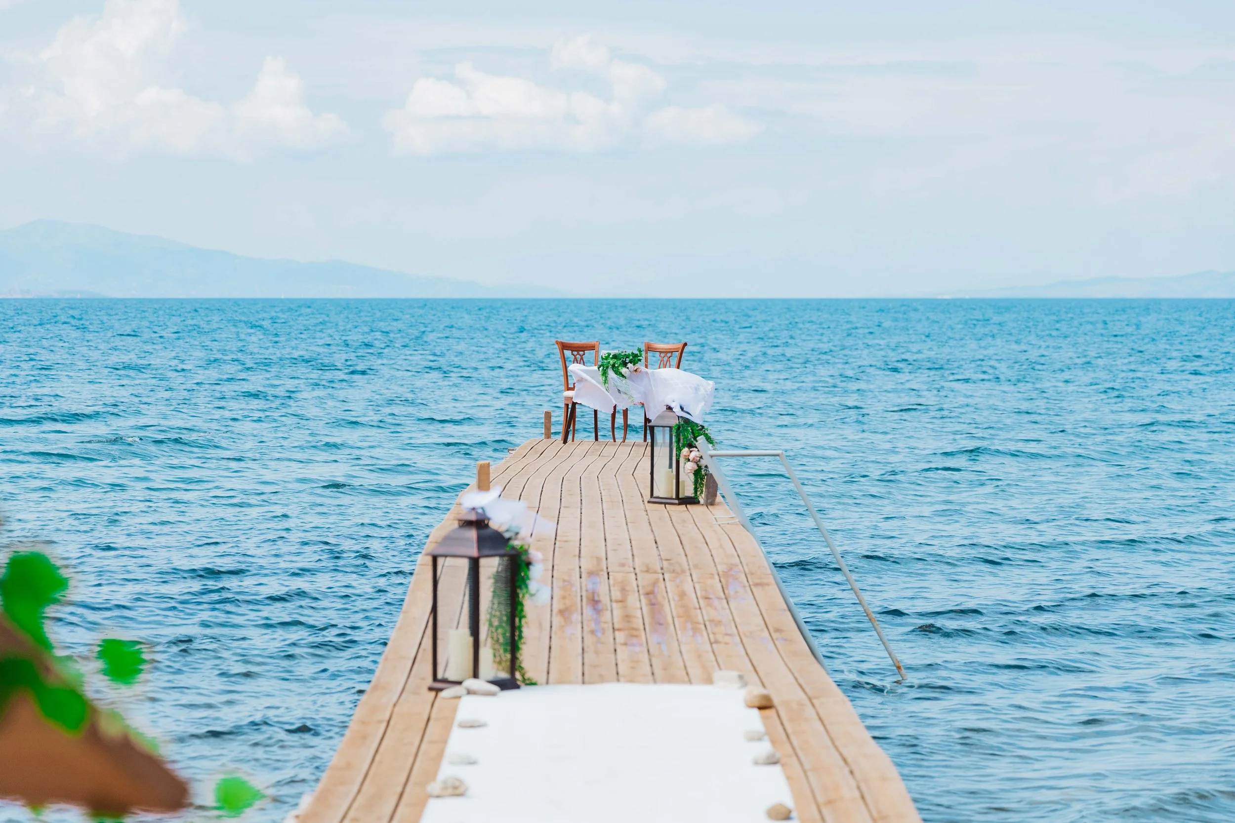 A wooden dock expands into a calm blue lake with mountains in the distance, decorated with lanterns, candles, and chairs for a wedding ceremony.