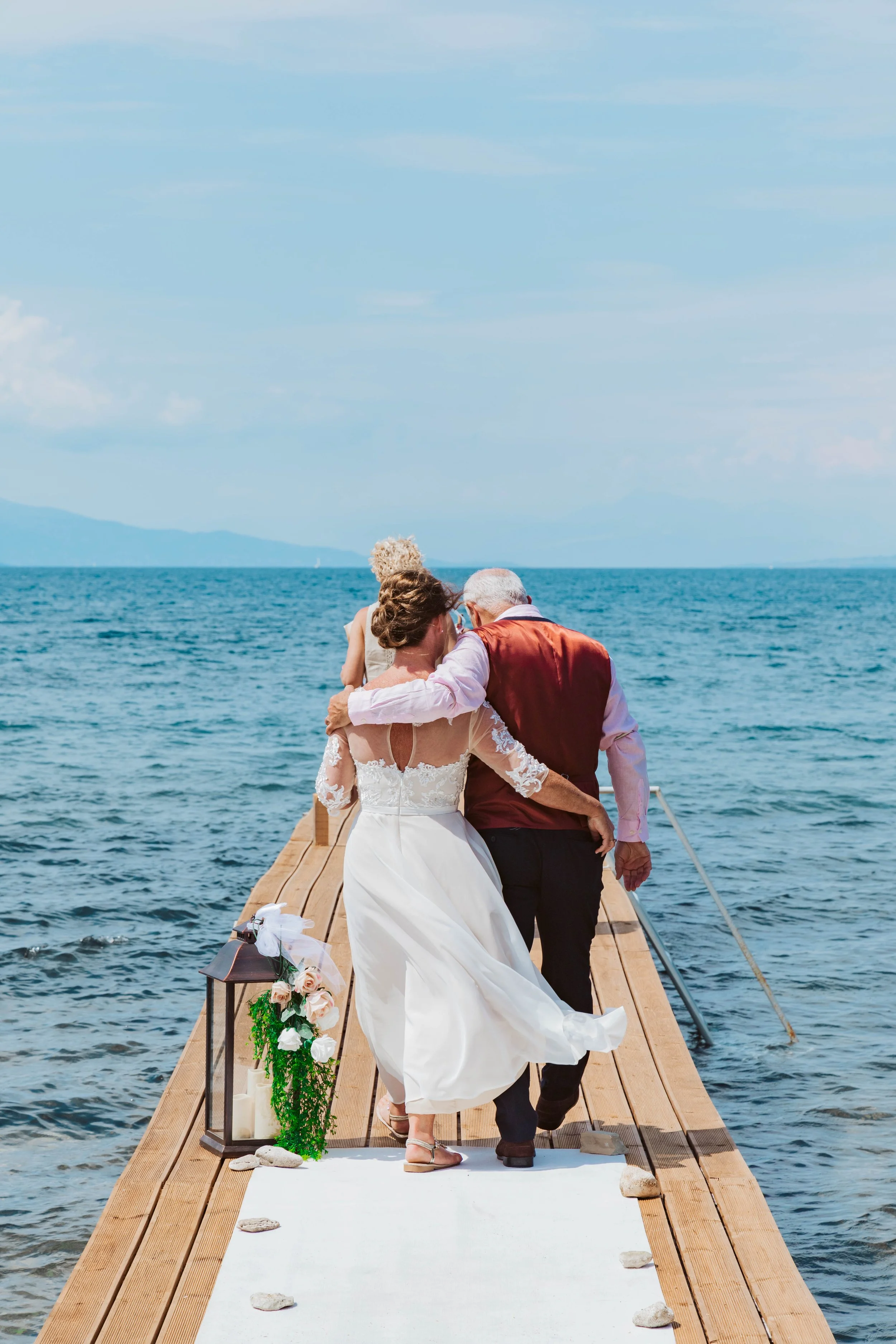 A couple walking on a wooden dock towards the water, with a woman in a white wedding dress and a man in a vest, accompanied by a child, a woman with curly blonde hair. Decorated lanterns and flowers are on the dock, and the setting is outdoors on a s