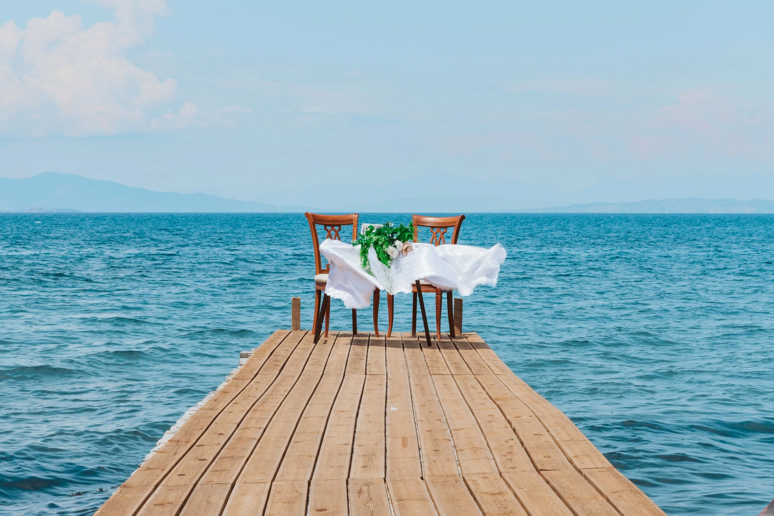 A wooden dock extends over the water with two chairs and a table set for a wedding or romantic event, decorated with a white cloth and a floral arrangement, overlooking a calm ocean with mountains in the background.