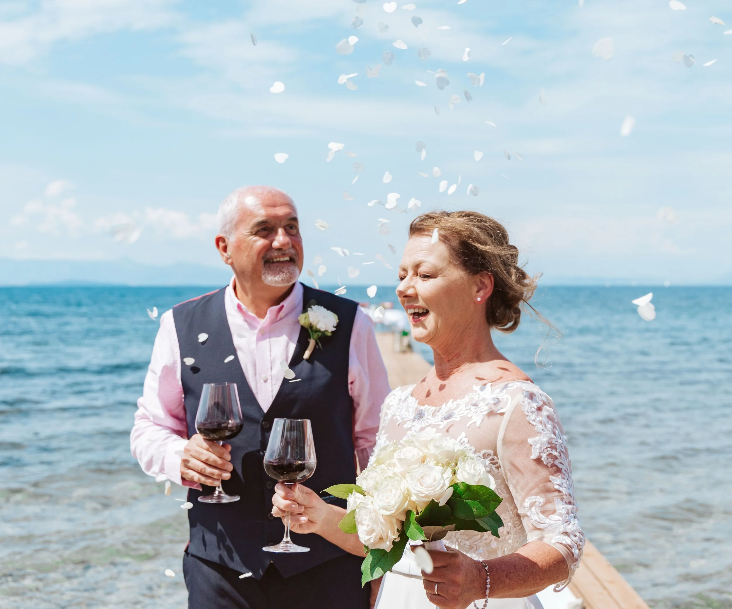 An elderly couple celebrating their wedding by the sea, holding glasses of red wine, with white confetti falling around them, and the bride holding a bouquet of white flowers.