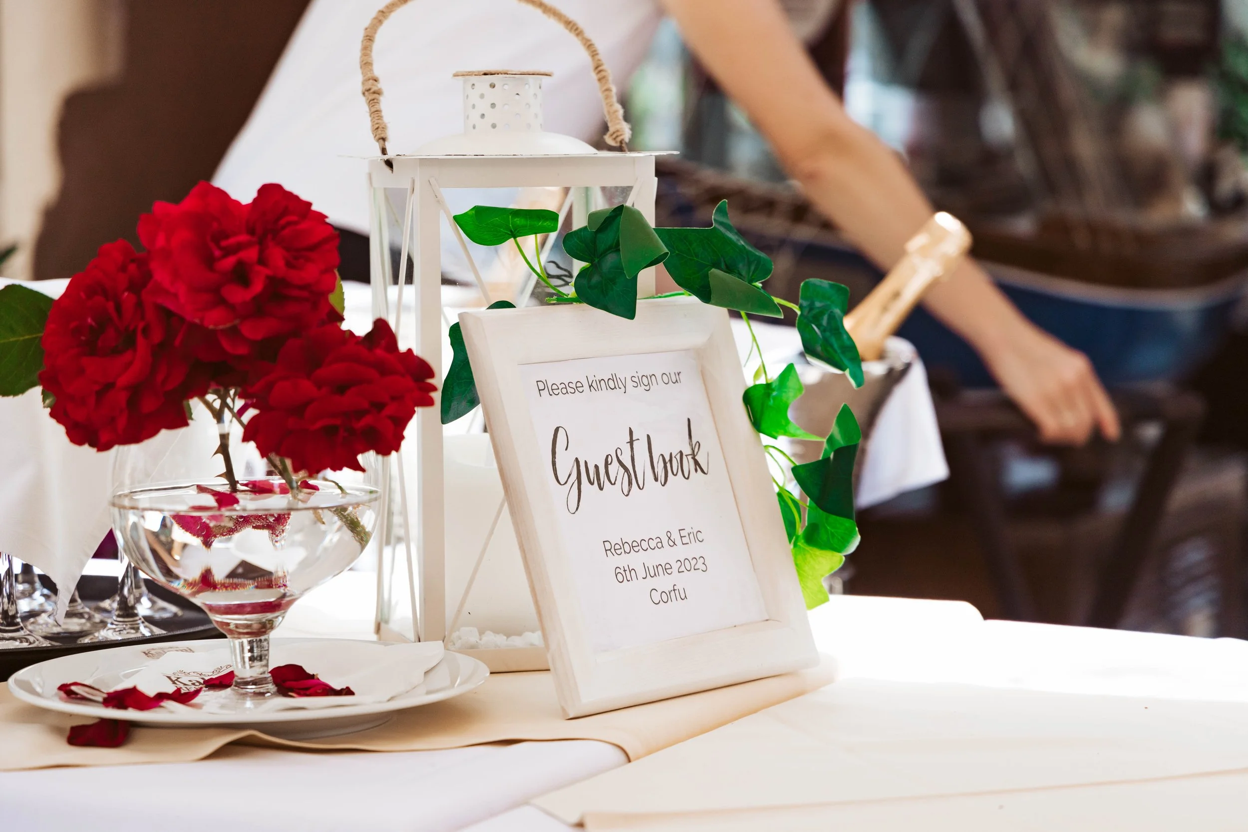 A guestbook sign at a wedding reception with red flowers, green leaves, and the names Rebecca & Eric, dated June 6th, 2023, in Corfu.