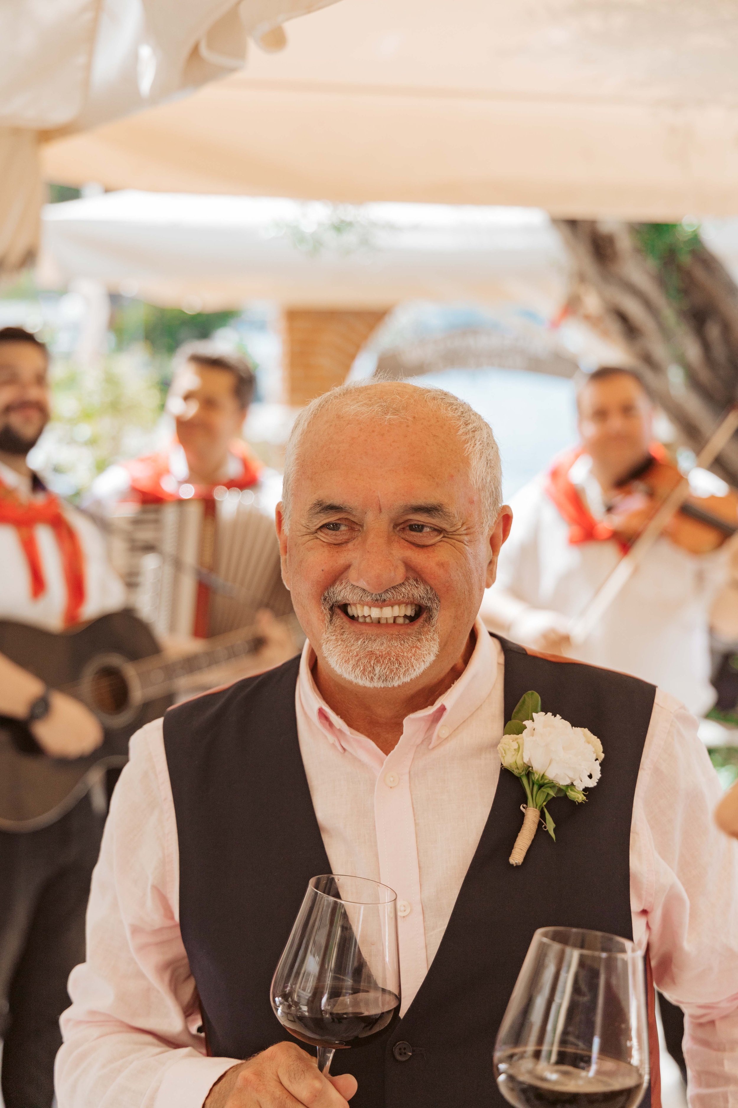 A smiling elderly man with a white beard and short hair, wearing a pink shirt with a black vest, holding a glass of red wine at a celebration. A corsage with white flowers is pinned to his vest. In the background, musicians playing guitars and accord