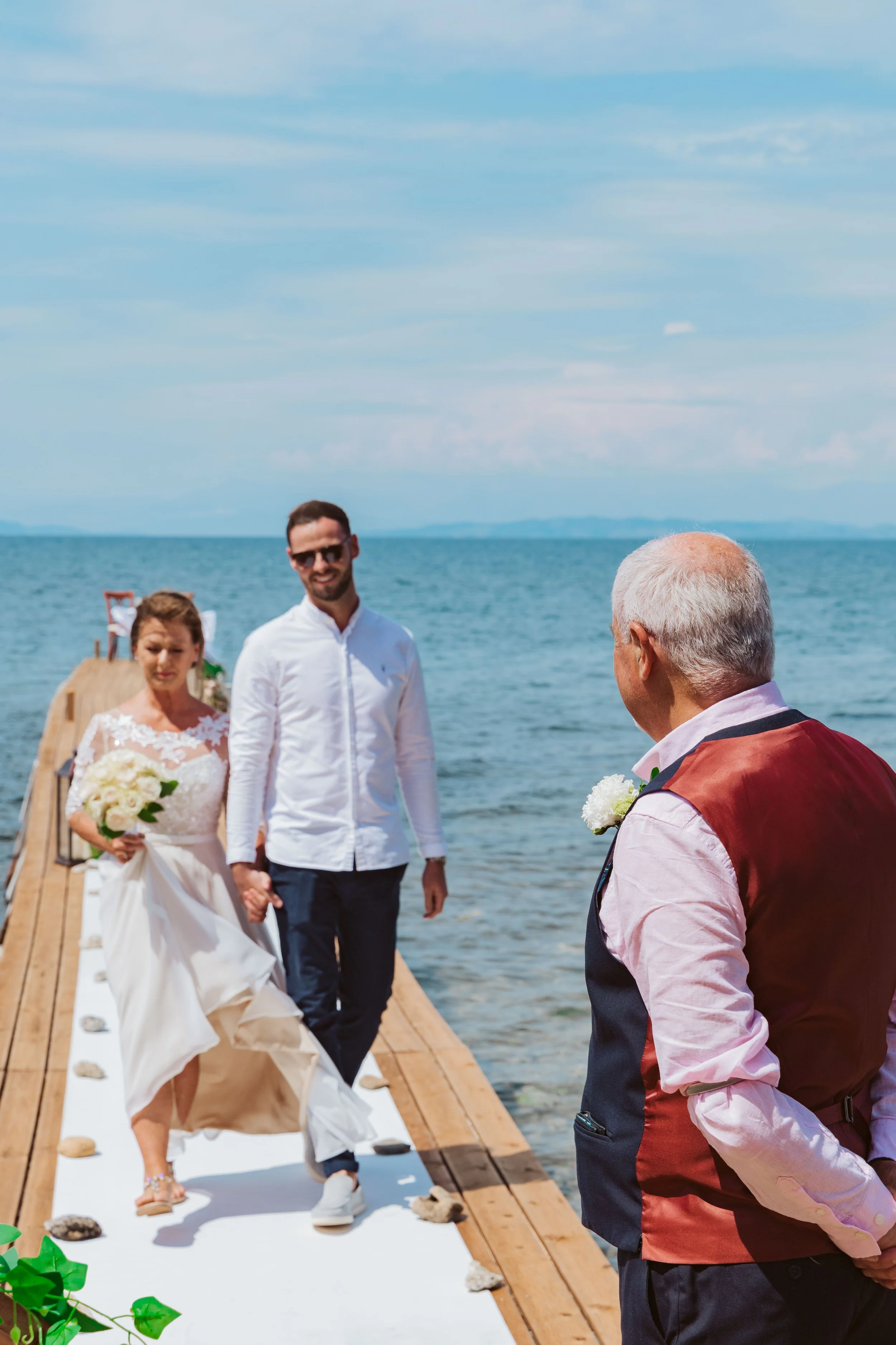 A bride and groom walking hand in hand on a wooden pier by the water, with an officiant standing nearby, during a wedding ceremony outdoors on a sunny day.