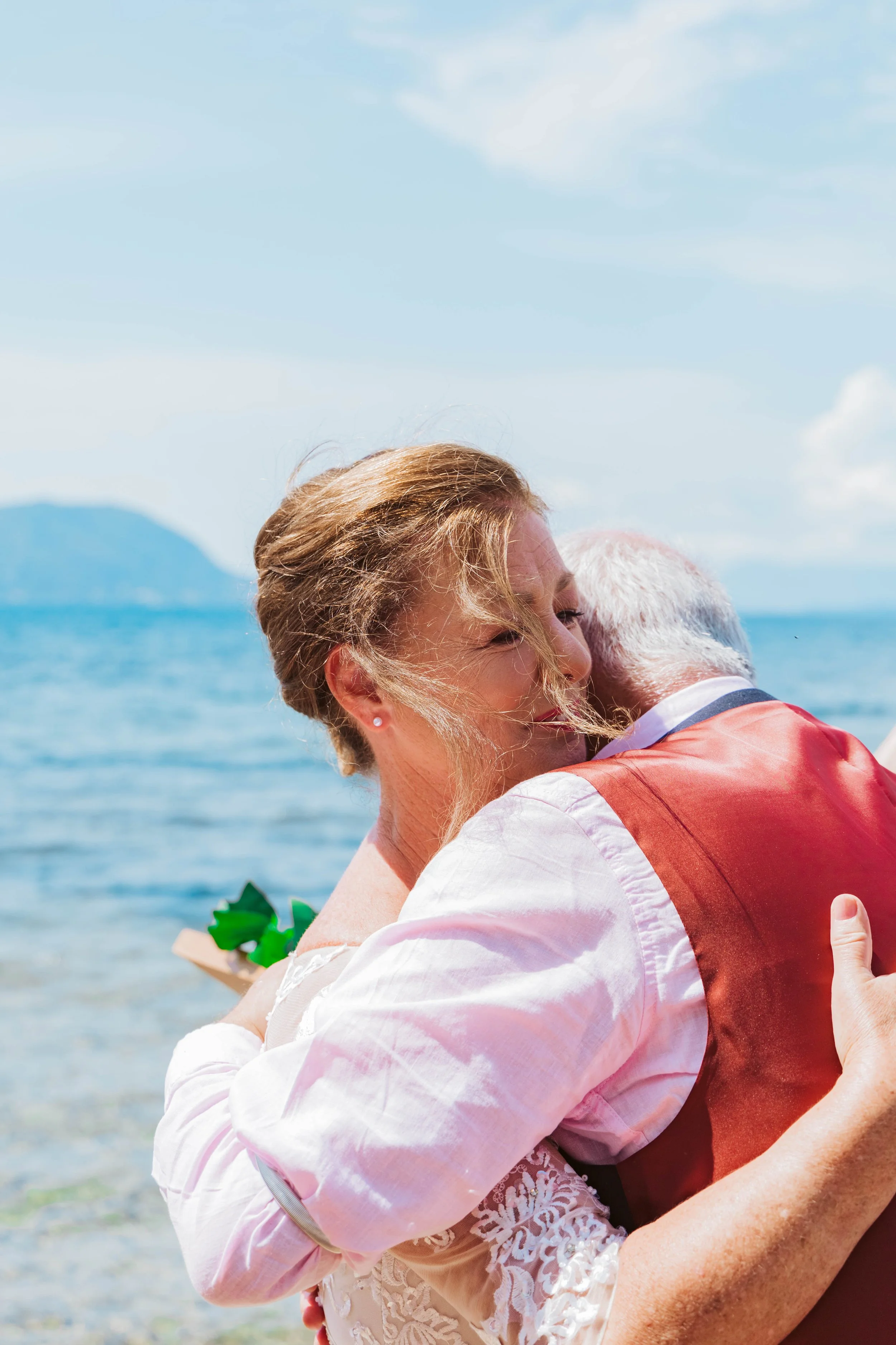 A woman and an elderly man hugging on a beach with a body of water and mountains in the background.