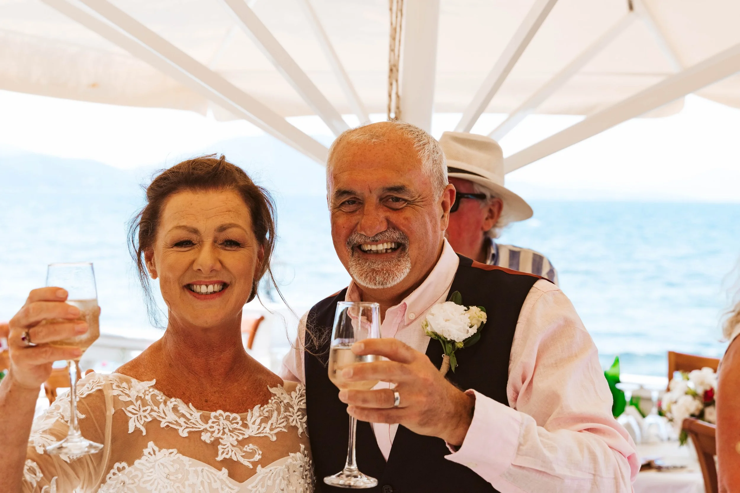 A smiling elderly couple raising glasses of champagne at a celebration near the ocean, under a white canopy, with a man wearing a striped shirt and a woman in a lace dress.