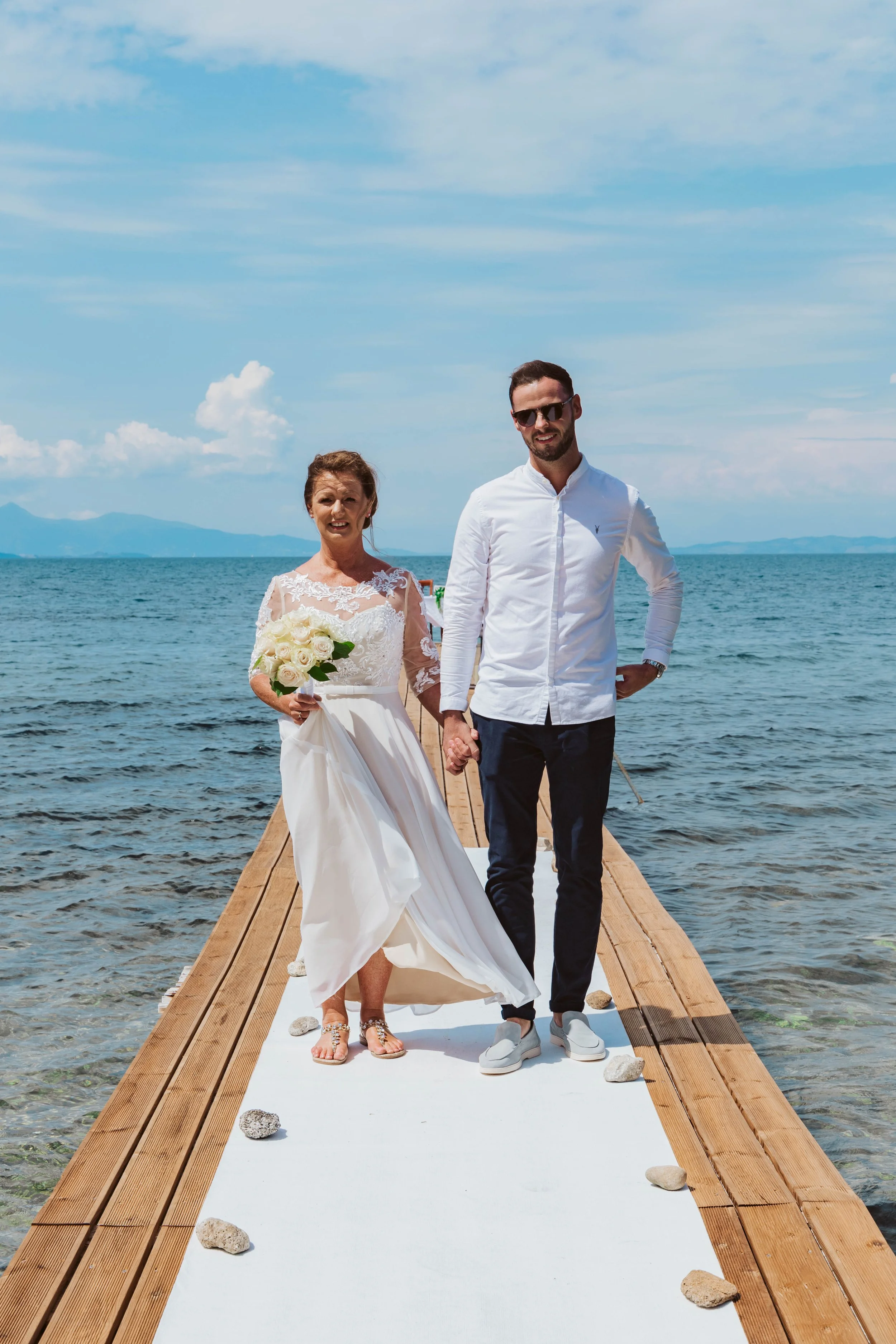 A bride and groom holding hands on a wooden dock over the water, with the bride in a white wedding dress holding a bouquet of white roses and the groom in a white shirt and dark pants, with mountains and blue sky in the background.