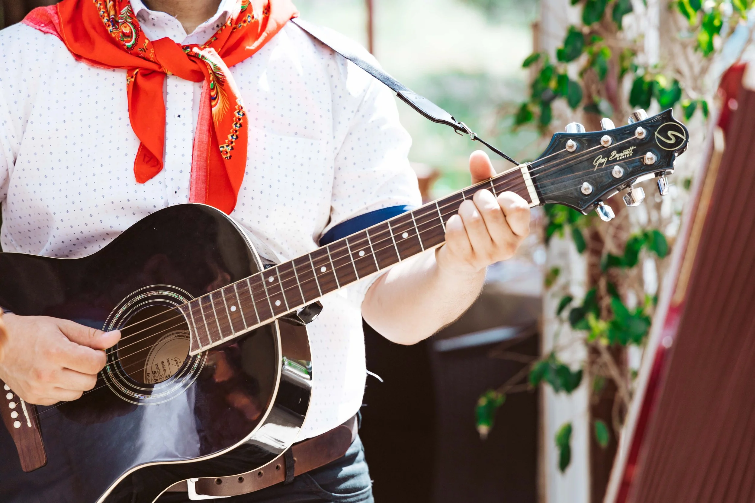Person playing a black acoustic guitar outdoors, wearing a white shirt with small dots, a red bandana, and a blue armband, with greenery and outdoor furniture in the background.