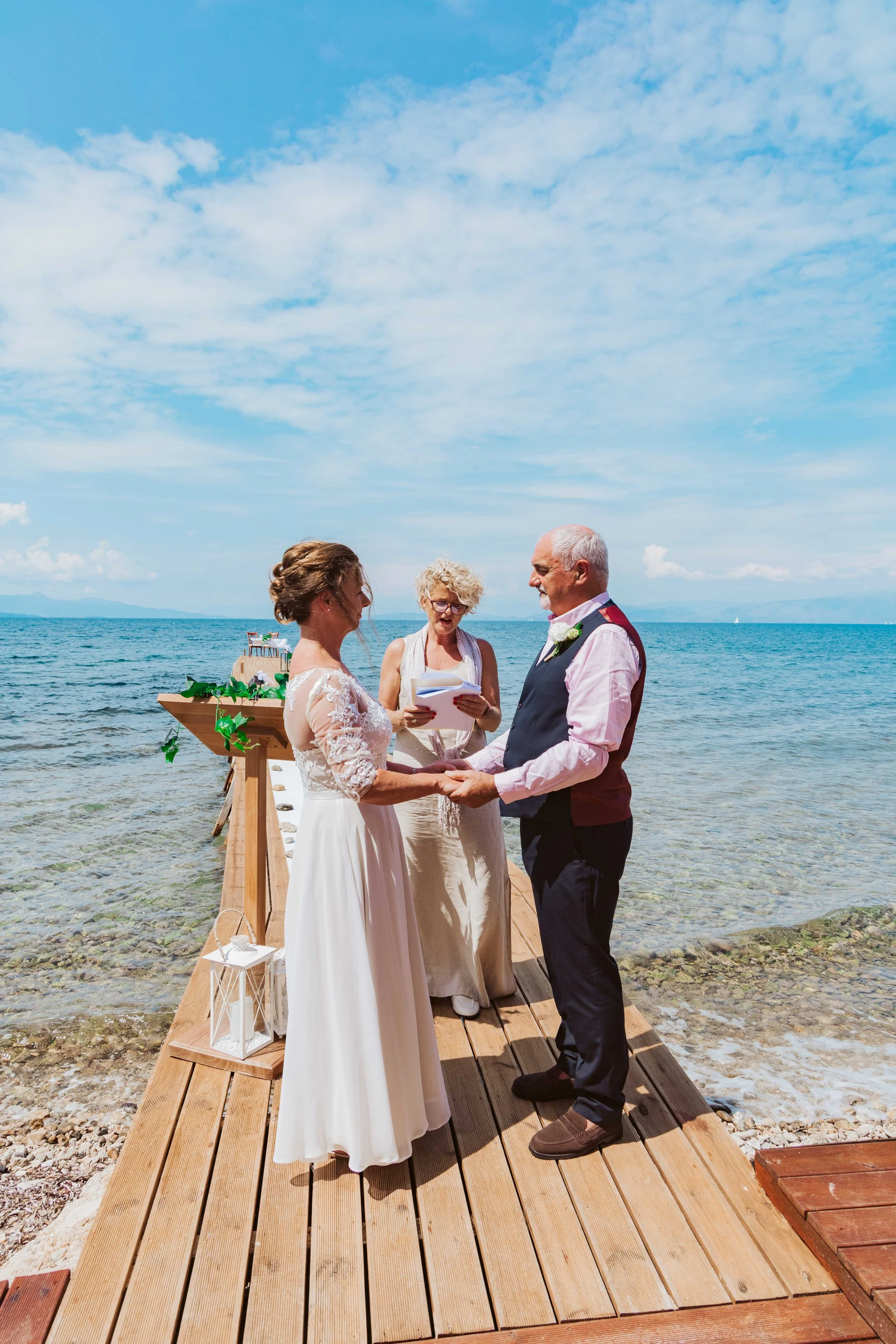 A couple getting married on a wooden dock by the water, with a person officiating and a scenic ocean background.
