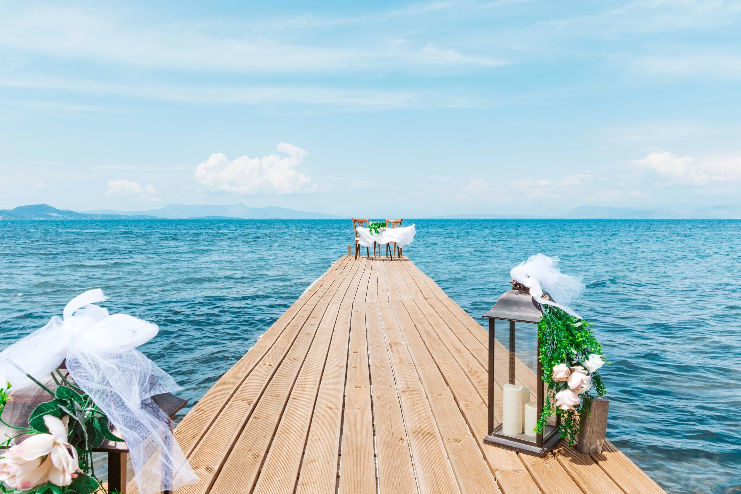 A wooden dock extending over the water with decorative lanterns, candles, and flowers, set for a wedding or special event by the ocean, with a distant landmass on the horizon under a blue sky with scattered clouds.