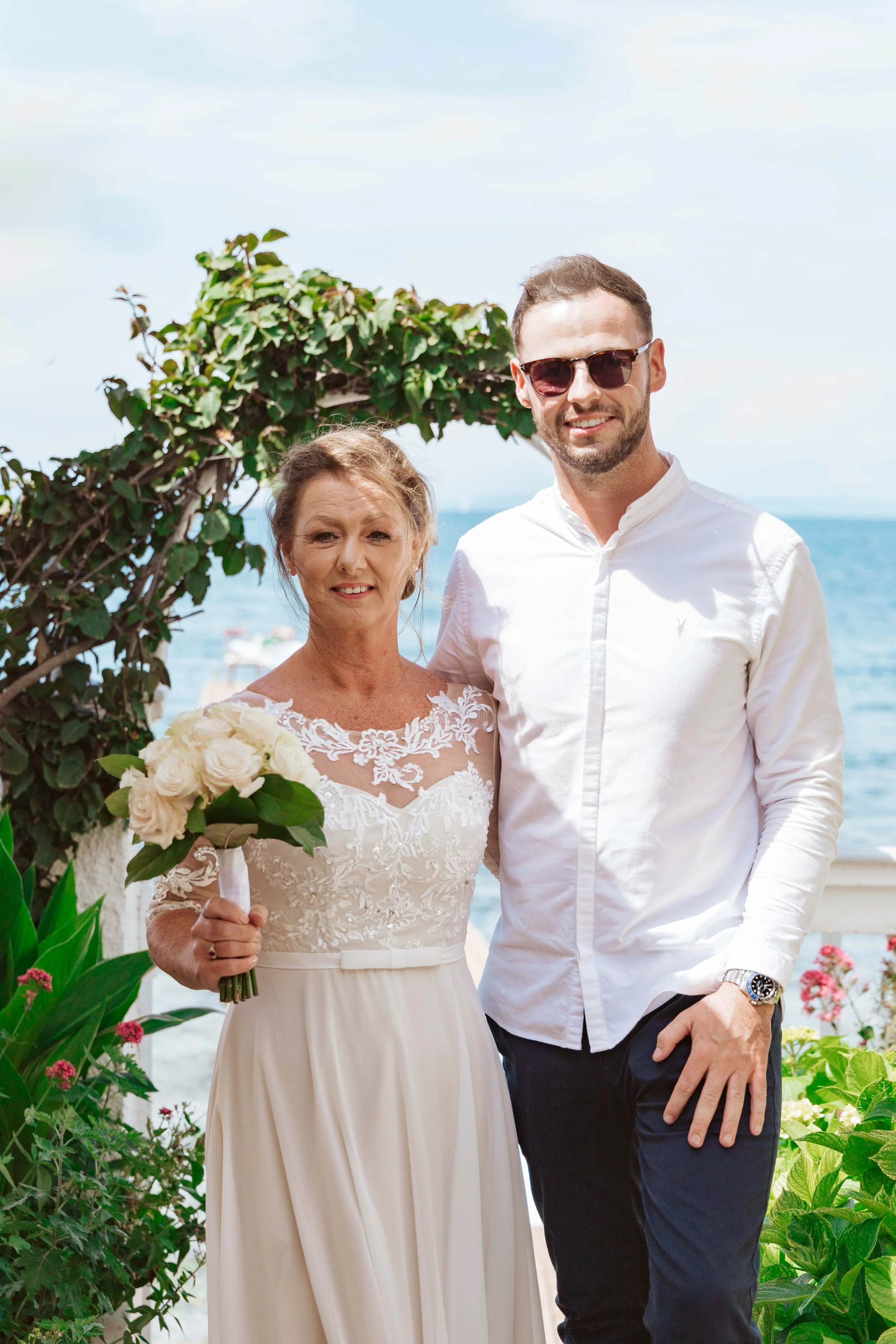 A woman in a white wedding dress holding a bouquet of white roses standing next to a man in sunglasses and a white shirt near the ocean, with greenery and a cloudy sky in the background.