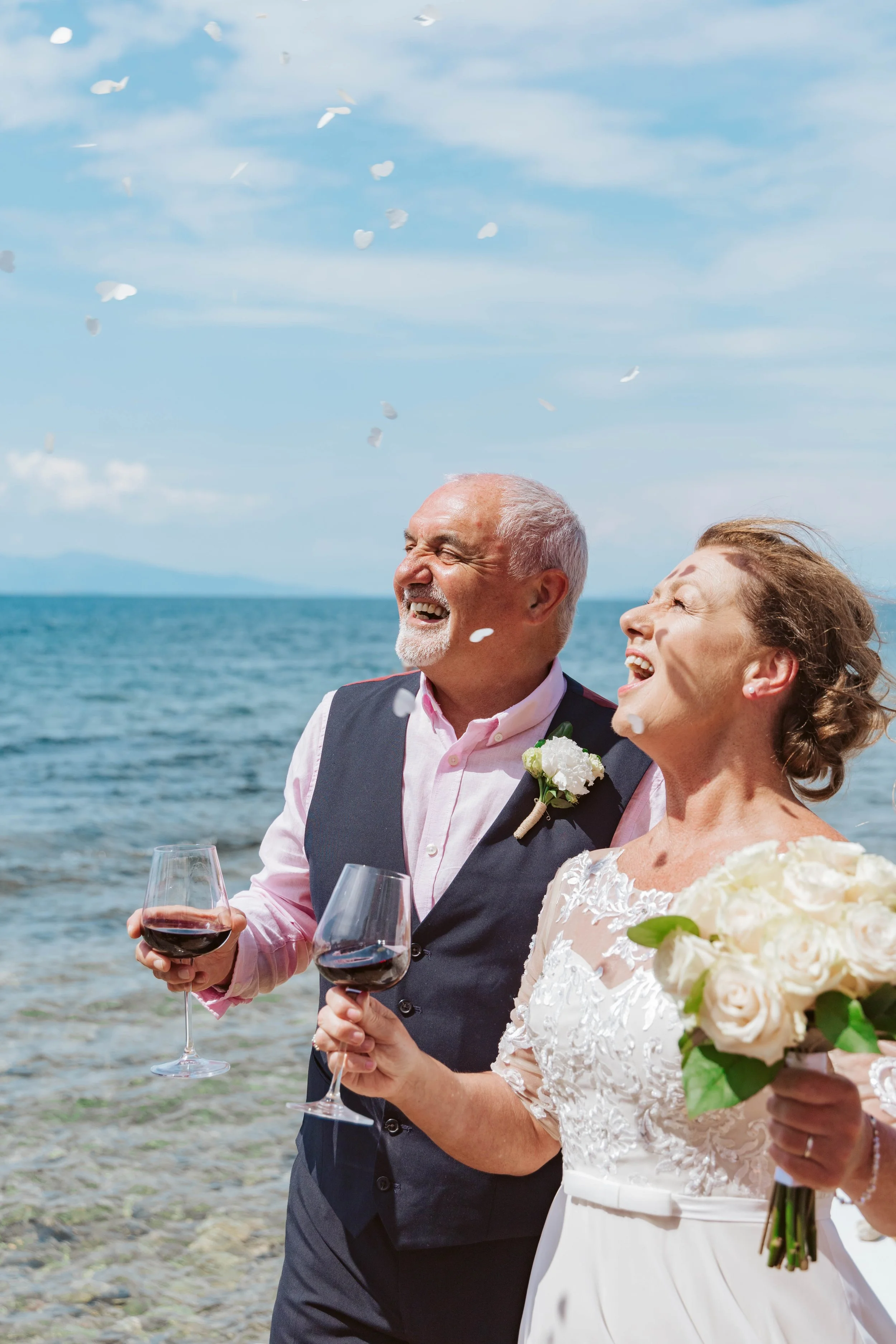 A happy elderly couple at a beach wedding, holding glasses of red wine, with a bouquet of white roses, smiling and celebrating.