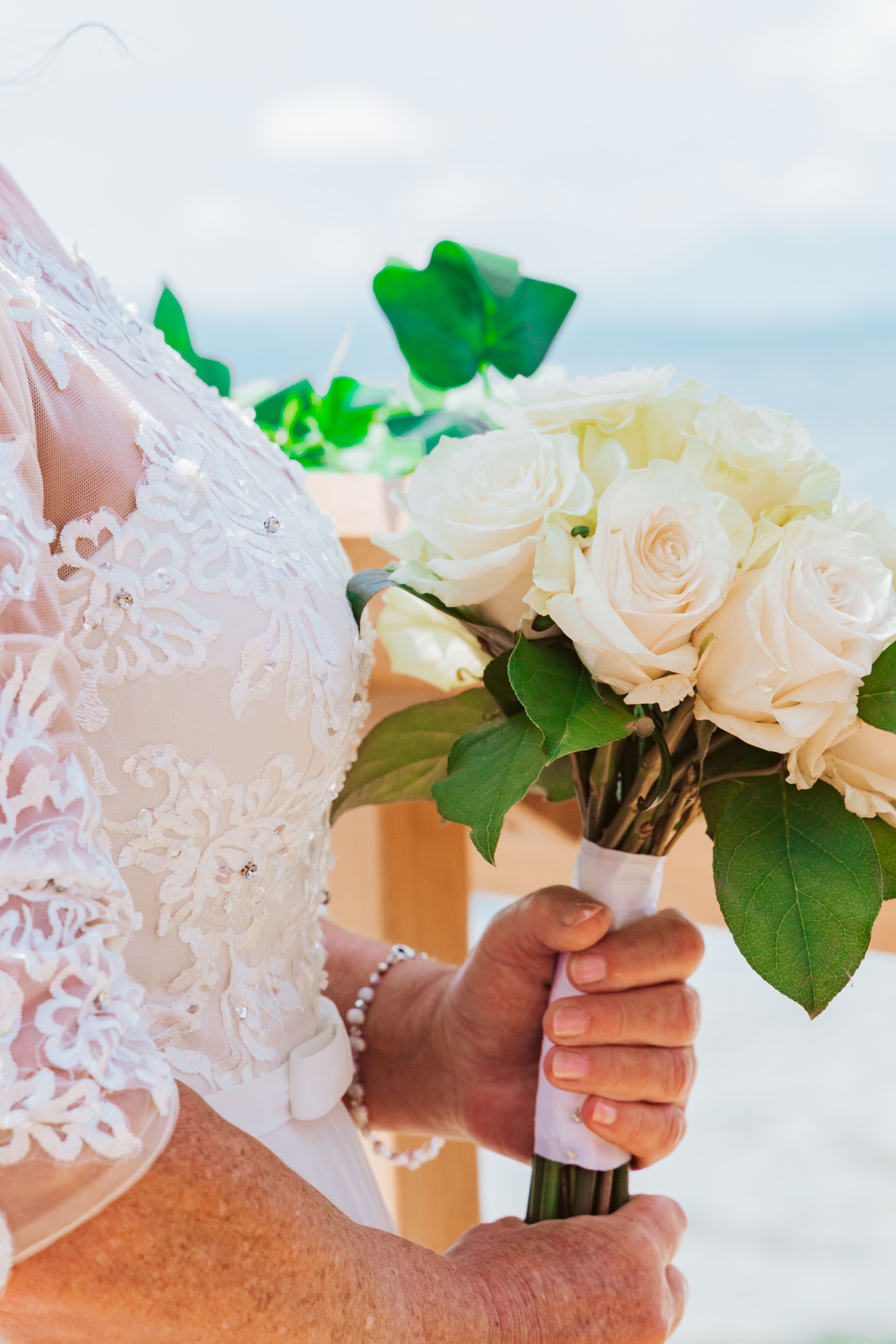 A close-up of a woman in a wedding dress holding a bouquet of white roses and green leaves.