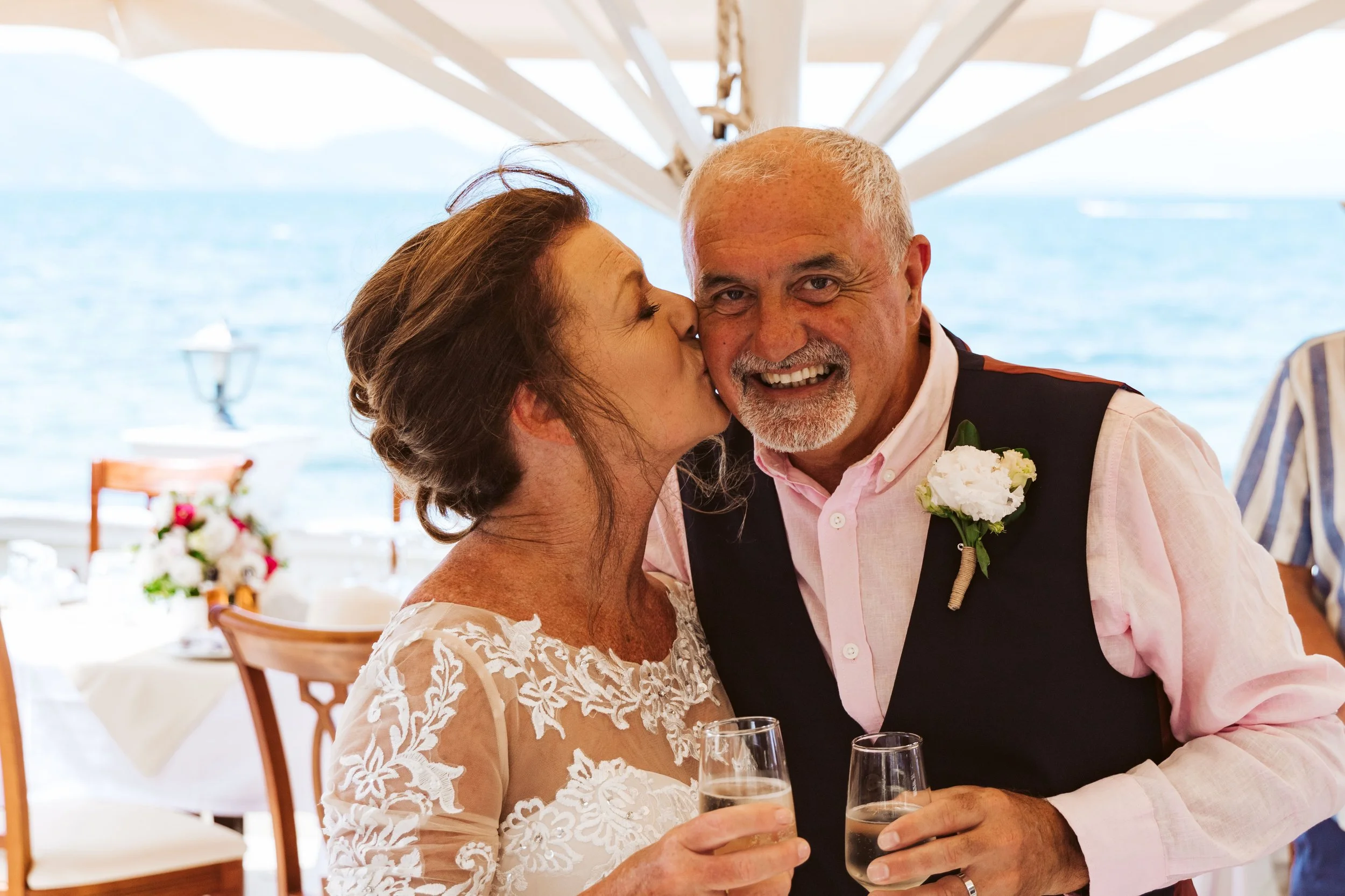 A woman kissing an older man on the cheek at a seaside wedding reception, holding a glass of champagne, with tables and chairs in the background overlooking the ocean.