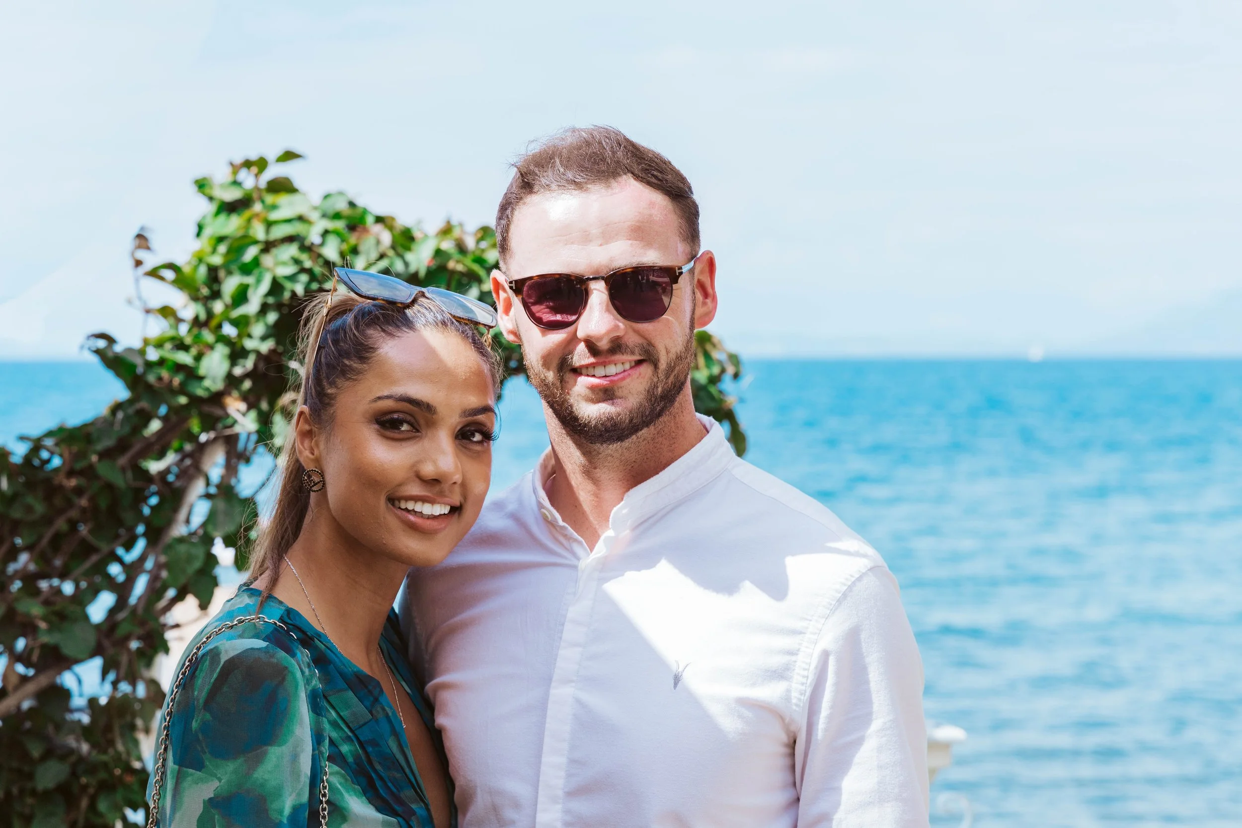 A smiling couple standing outdoors near the ocean, with a bush and blue water in the background. The woman has sunglasses on her head and is wearing a patterned top, and the man is wearing sunglasses and a white shirt.