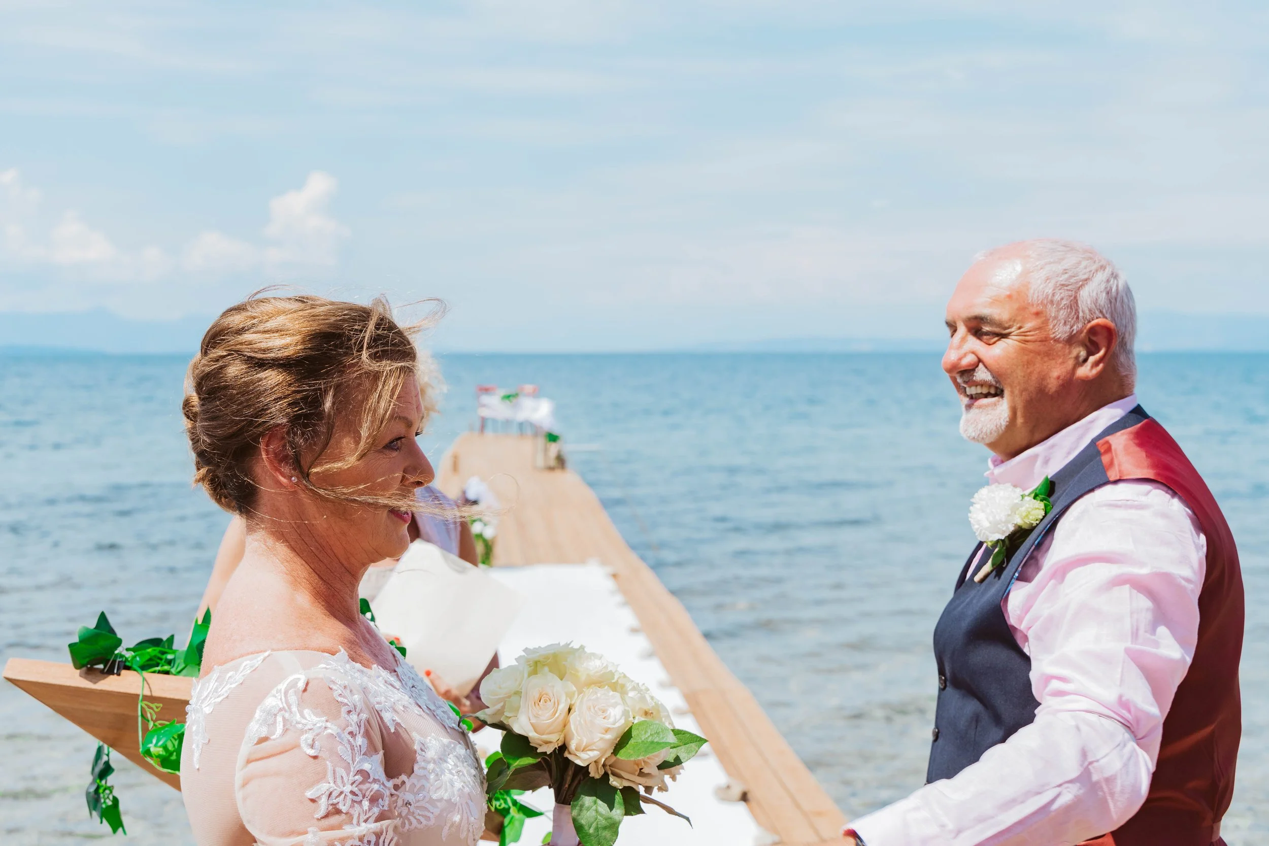 A bride and groom smiling at each other during their wedding ceremony on a beach, with an ocean and a wooden aisle in the background.