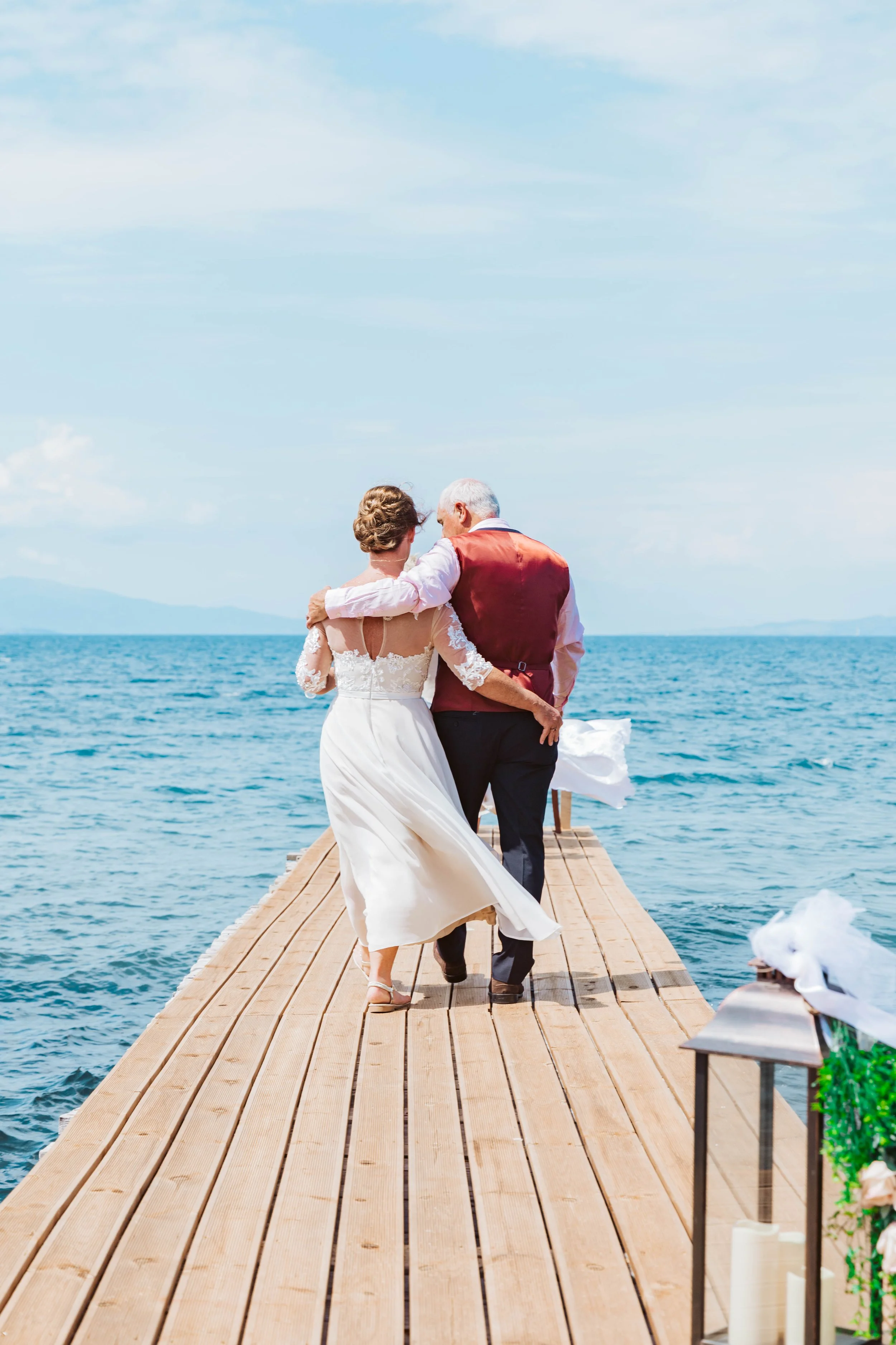 A bride and groom walking arm-in-arm on a wooden pier over the water, dressed in wedding attire, with the bride in a white dress and the groom in a vest and dress pants, as they enjoy a romantic moment by the sea.
