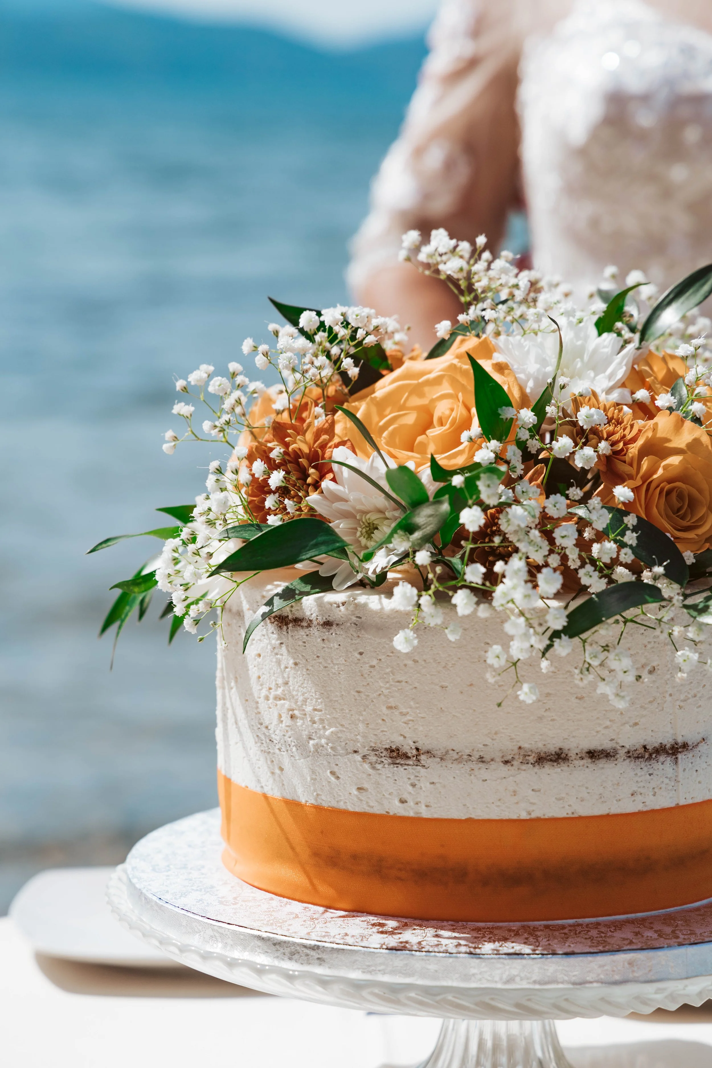 A wedding cake decorated with orange and white flowers, greenery, and an orange ribbon, set against a beach background.
