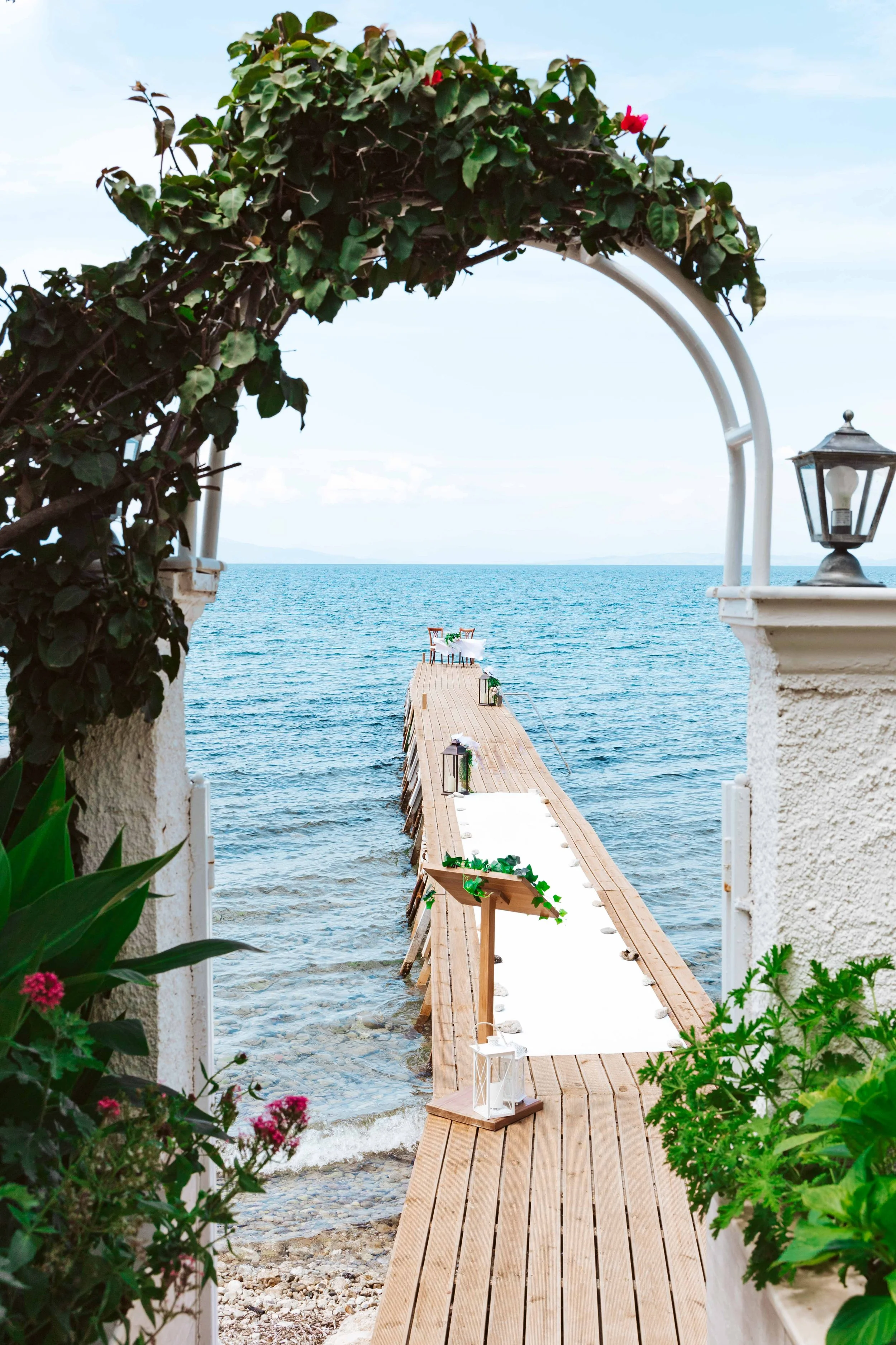 A scenic wooden pier extending into the ocean, viewed through an archway decorated with green ivy and pink flowers, with lanterns on the sides of the pier, leading to a table set for an outdoor event by the water.