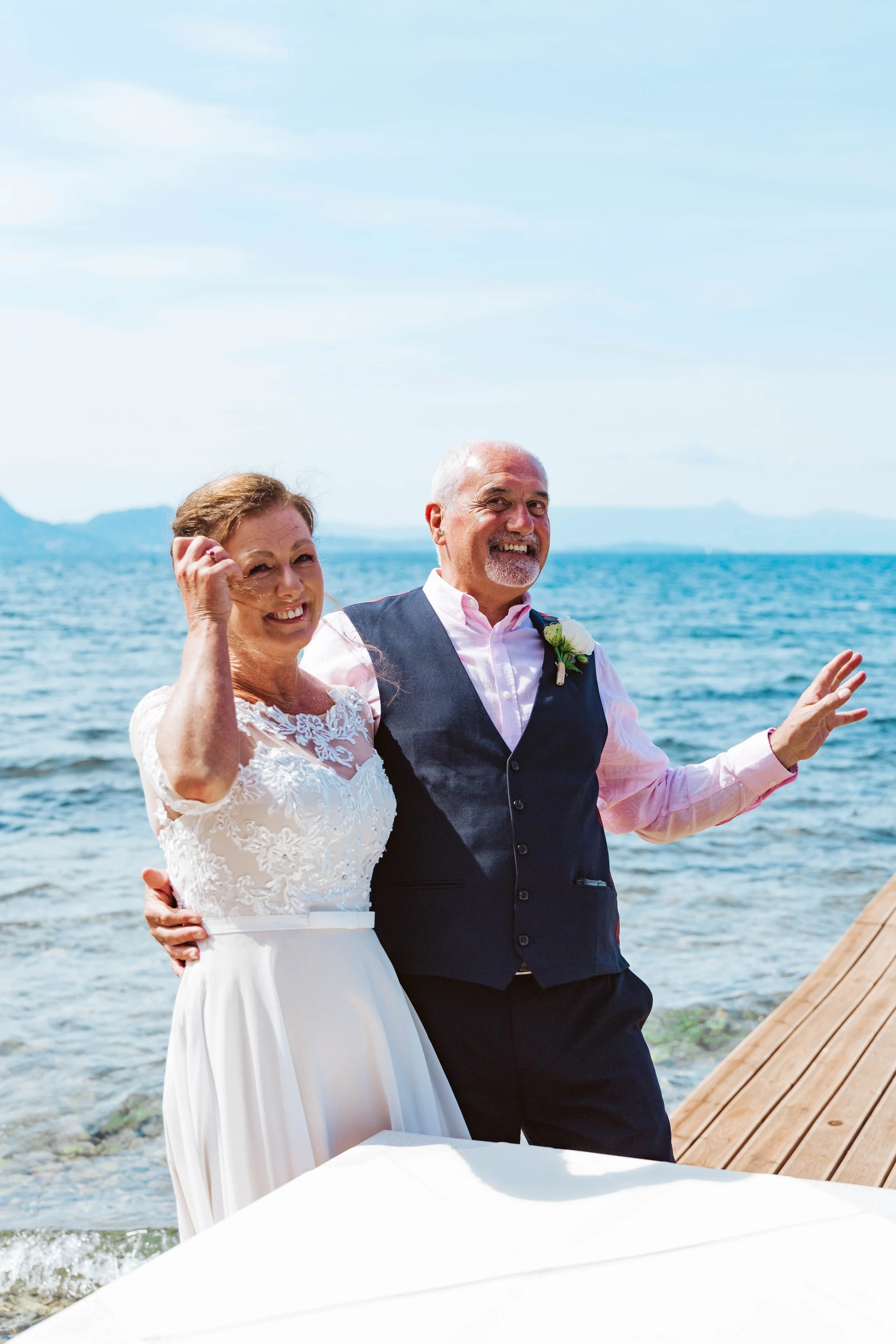 A smiling older bride and groom standing on a dock by the water, celebrating their wedding outdoors on a sunny day.