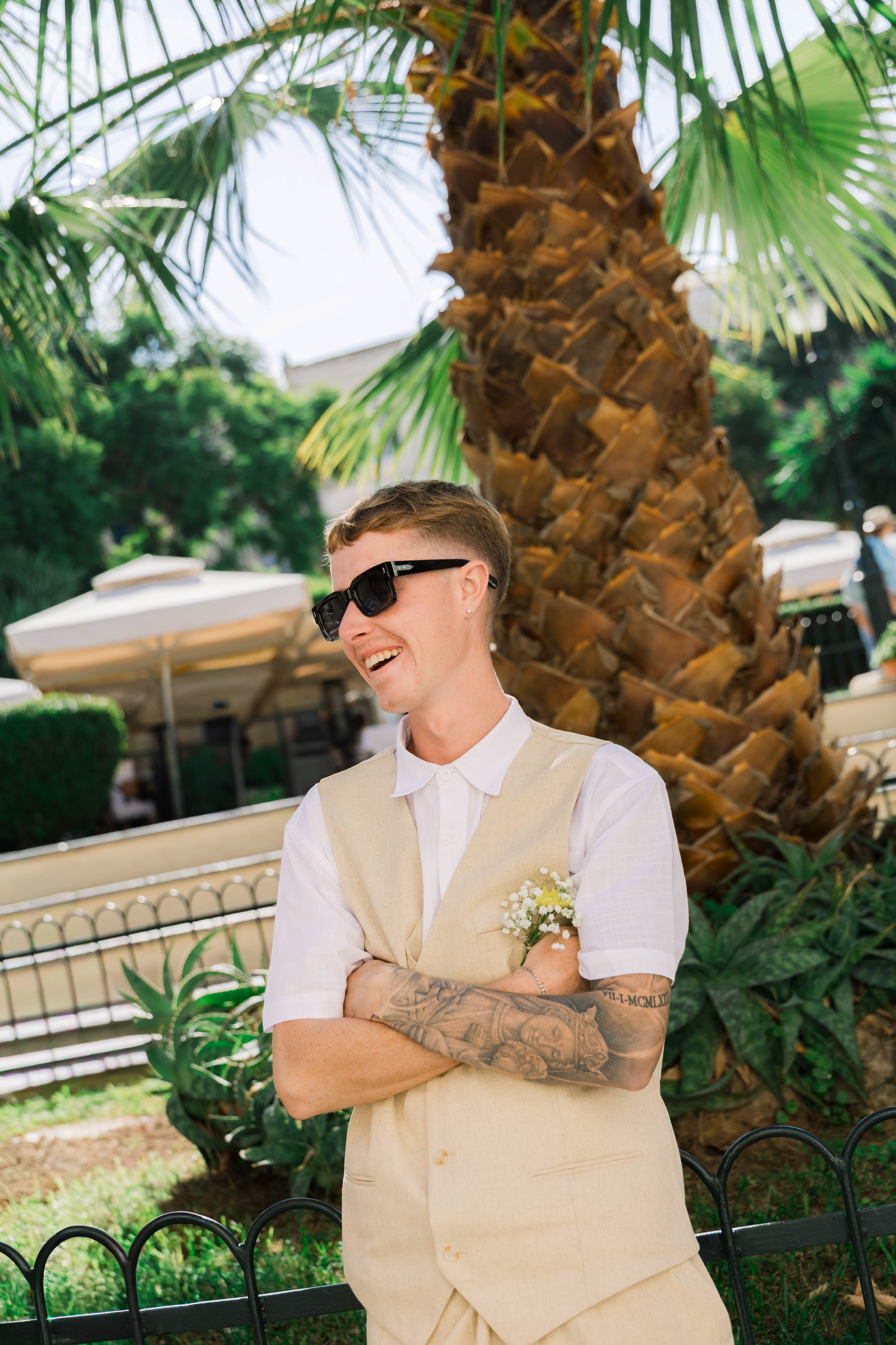 A man with short hair, sunglasses, a white shirt, and a light beige vest, smiling outdoors near a palm tree. He has tattoos on his left arm and a flower boutonniere on his vest. The background includes benches, umbrellas, and greenery.
