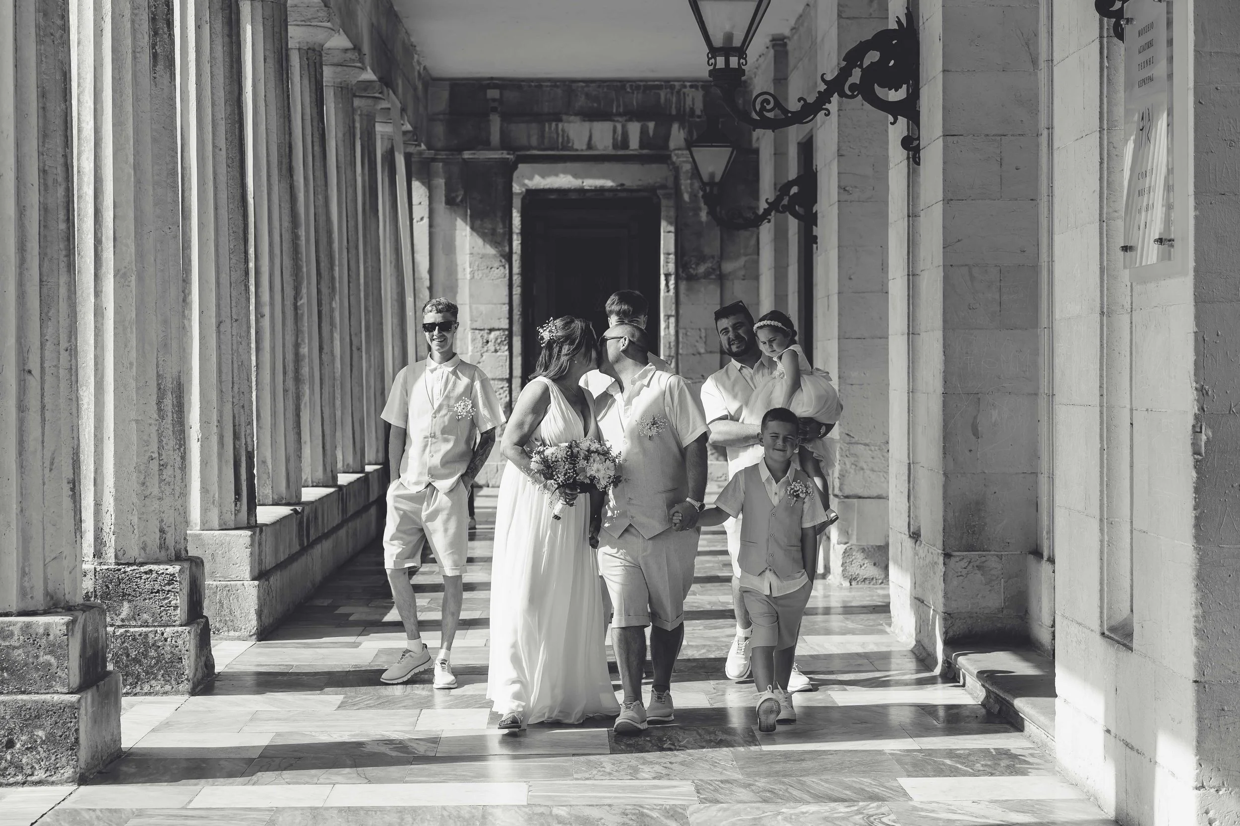 A black-and-white photo of six people, including a bride, groom, children, and guests, walking through a colonnaded walkway outside a stone building, likely at a wedding.