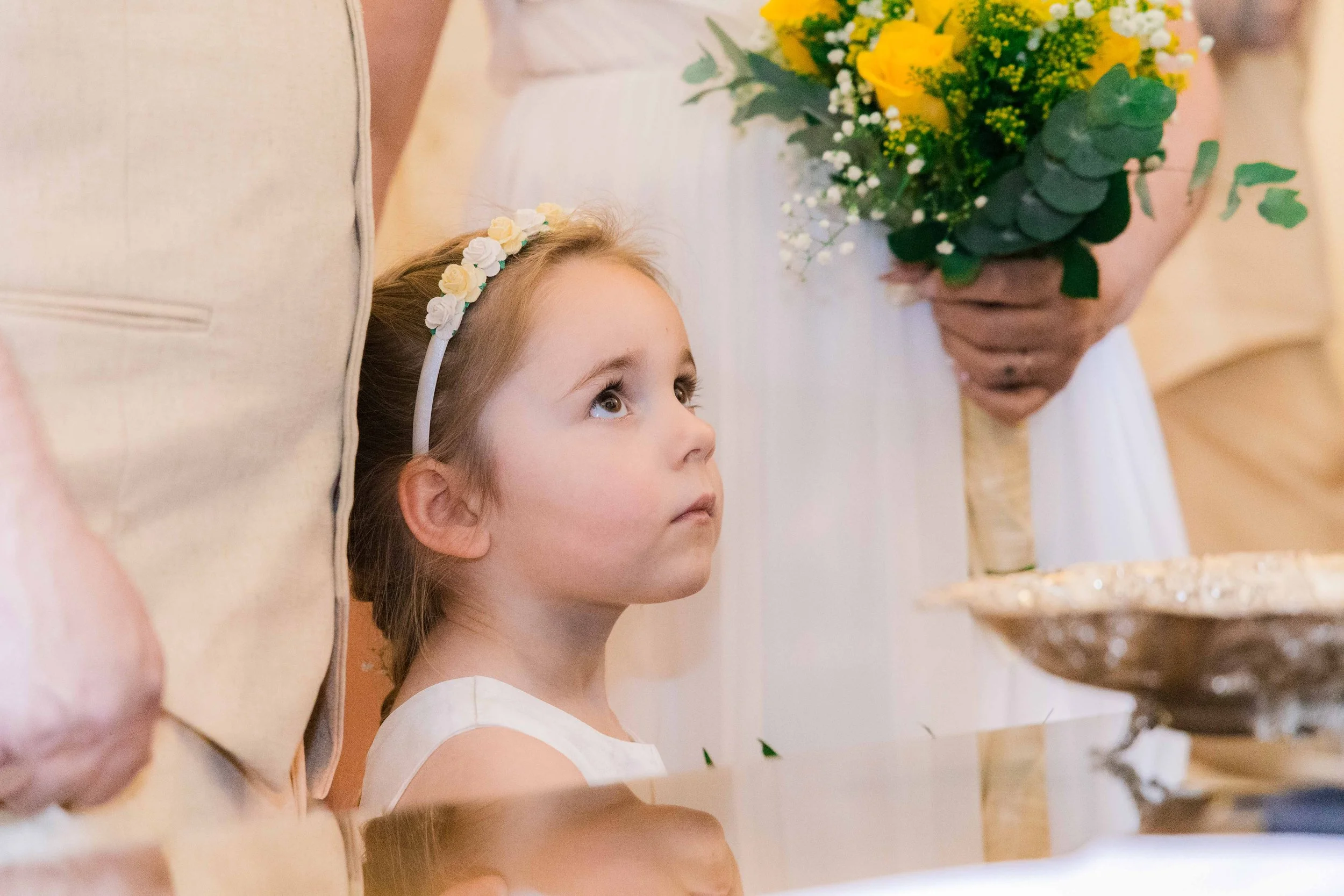 Young girl with a floral headband looking up at an adult holding a bouquet of yellow and white flowers during a wedding ceremony.