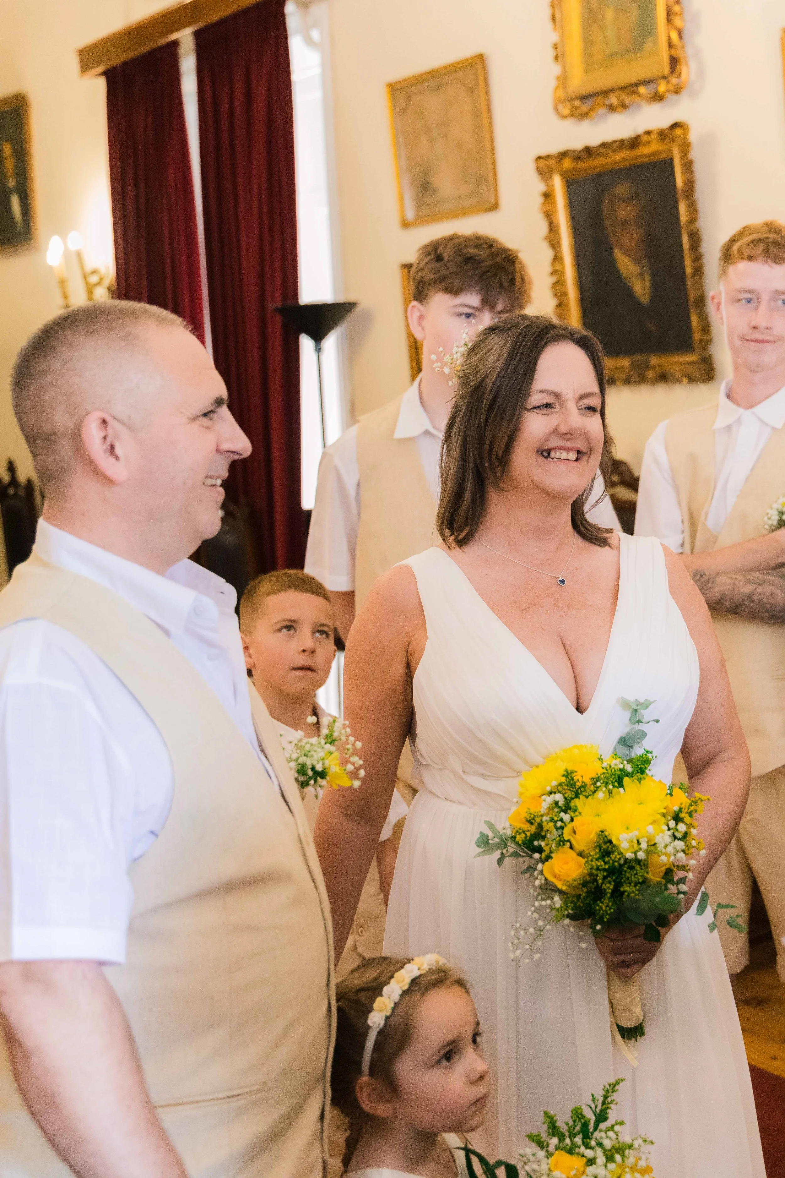 A woman in a white dress holding a bouquet of yellow flowers, smiling with others in a room decorated with paintings, red curtains, and vintage furniture.
