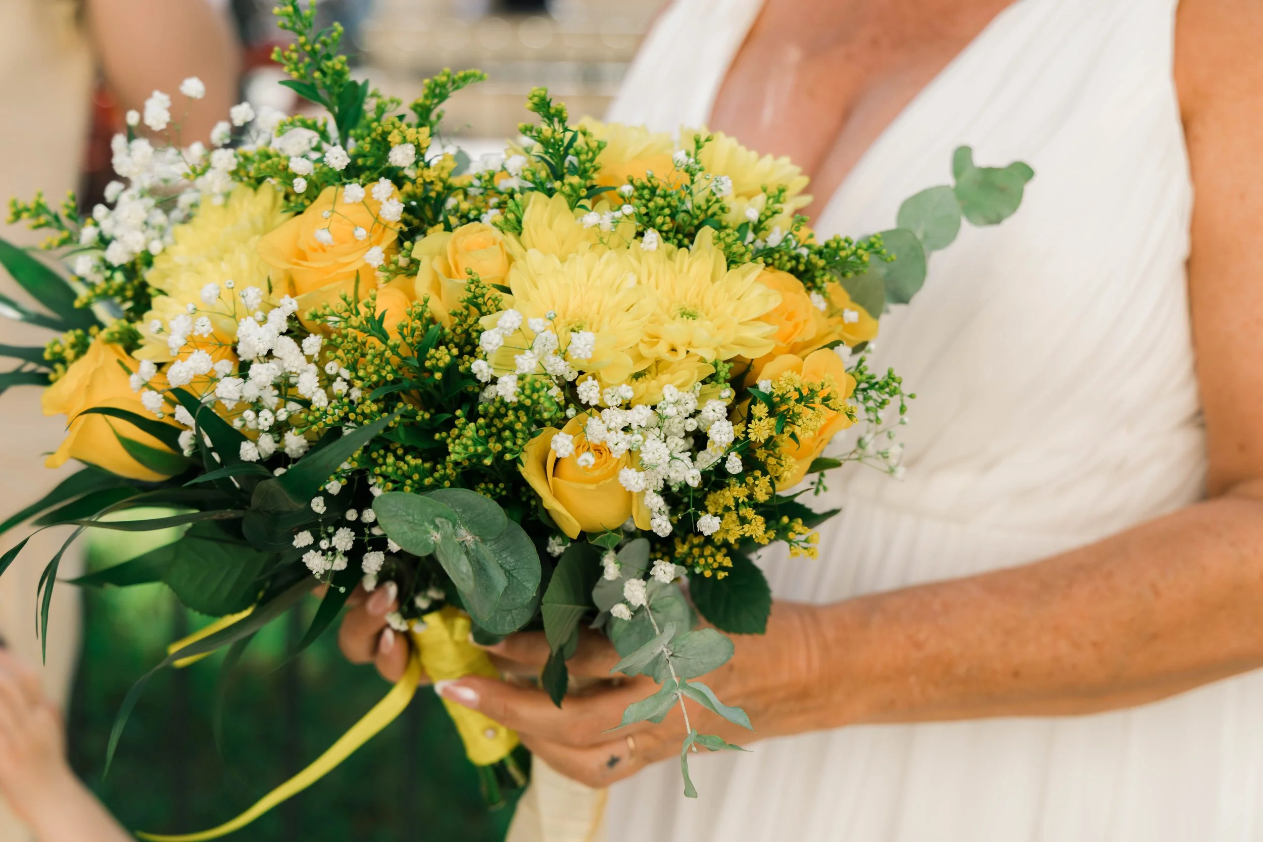 A woman in a white dress holding a yellow floral bouquet with roses, daisies, baby's breath, and greenery.