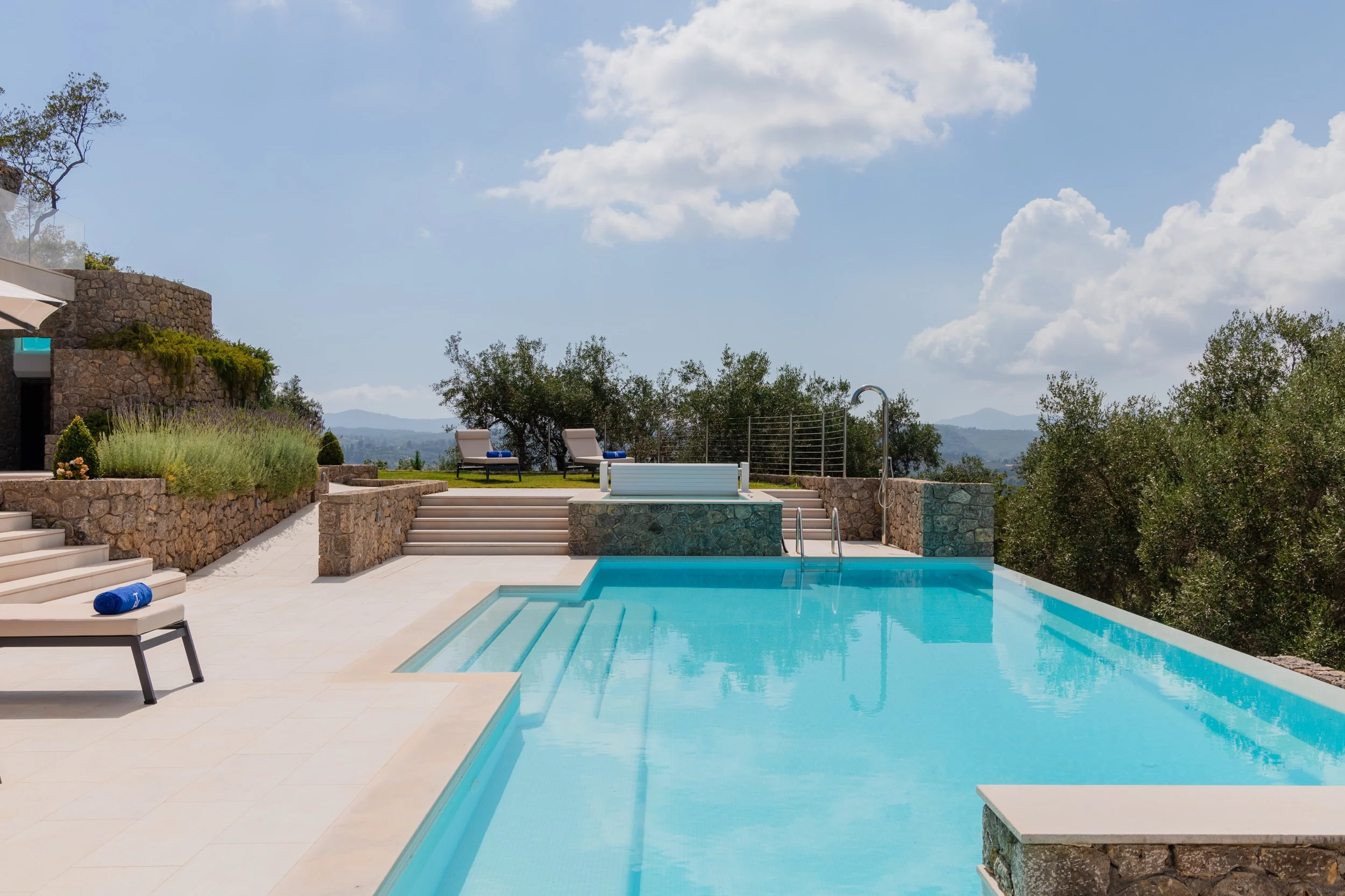 Luxury pool area with steps, lounge chairs, greenery, and a view of distant mountains under a partly cloudy sky.