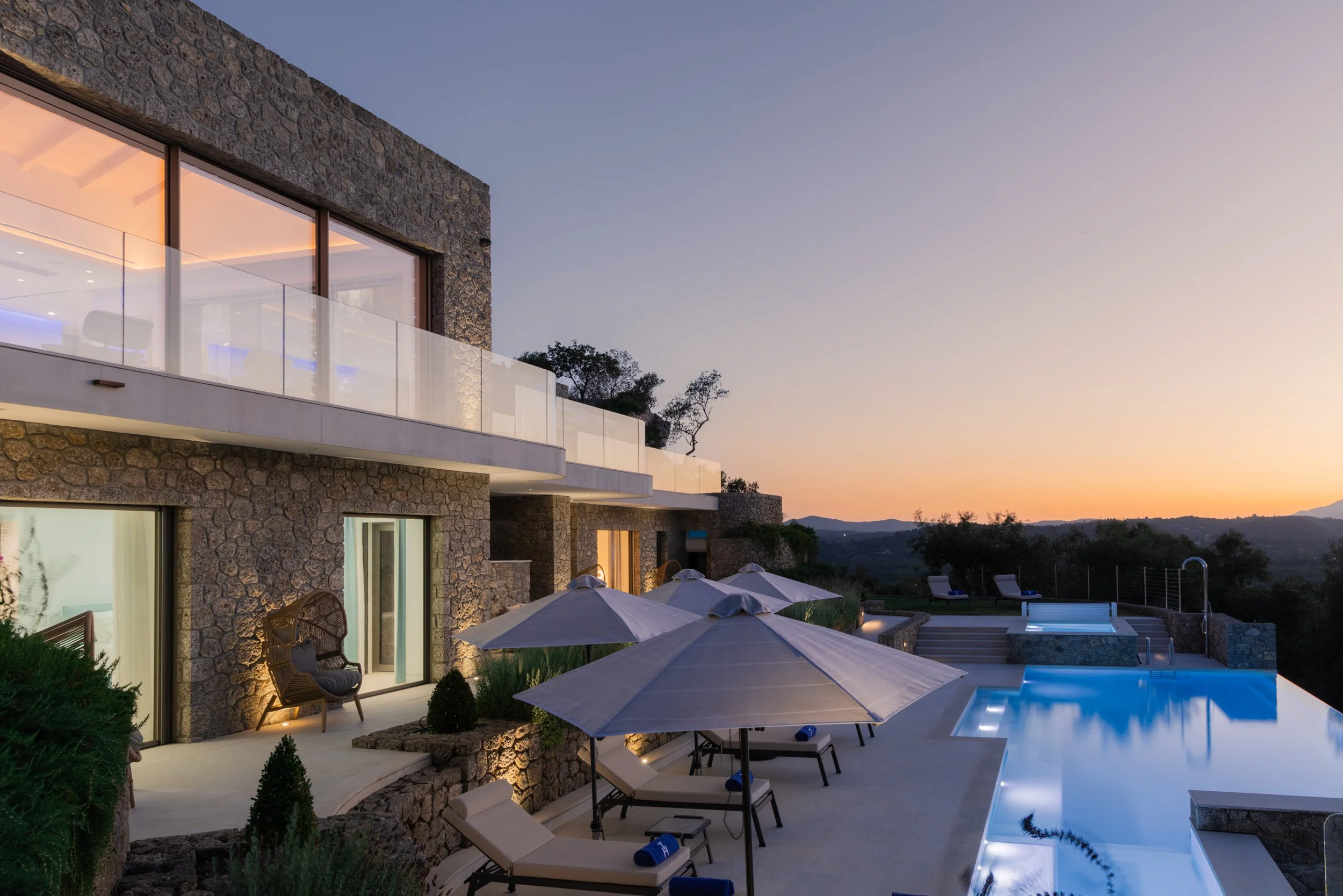 Luxury house with stone exterior, glass balcony, outdoor pool, and lounge chairs with umbrellas at sunset.