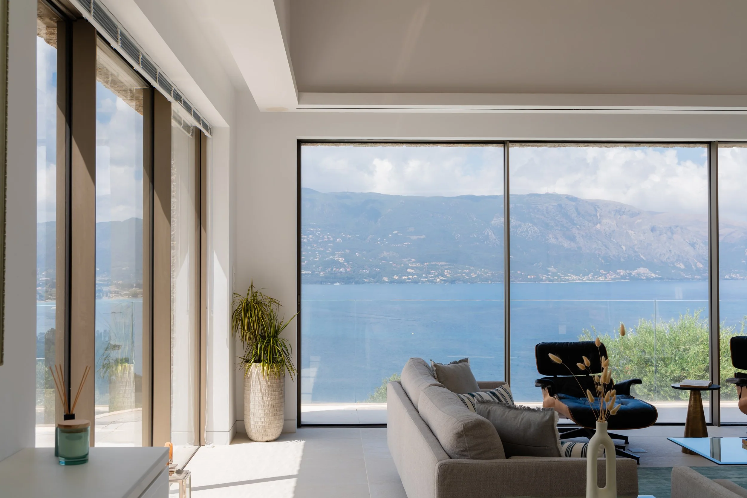 Modern living room with large windows overlooking a lake and mountains, featuring a white sofa, black lounge chair, potted plant, and decorative items.