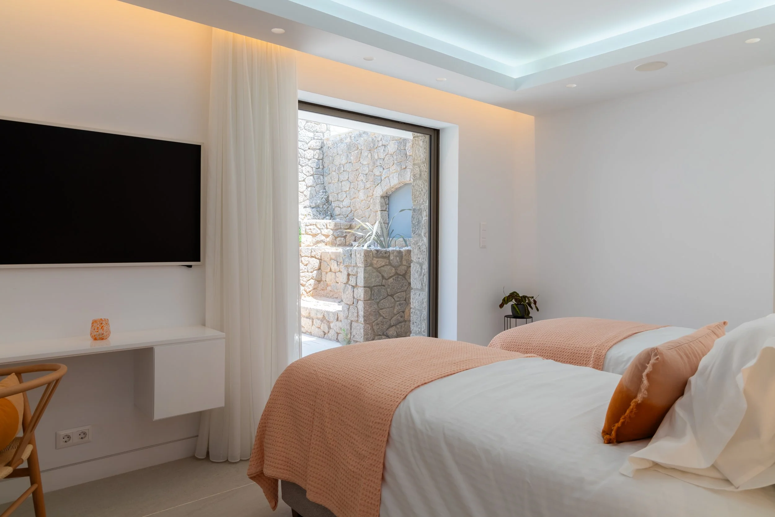 Modern bedroom with two beds, pink blankets, white pillows, a large window with white curtains, a doorway leading outside, a flat-screen TV on the wall, a small desk with a daily-themed candle, a small side table with a plant, and a ceiling with skyl