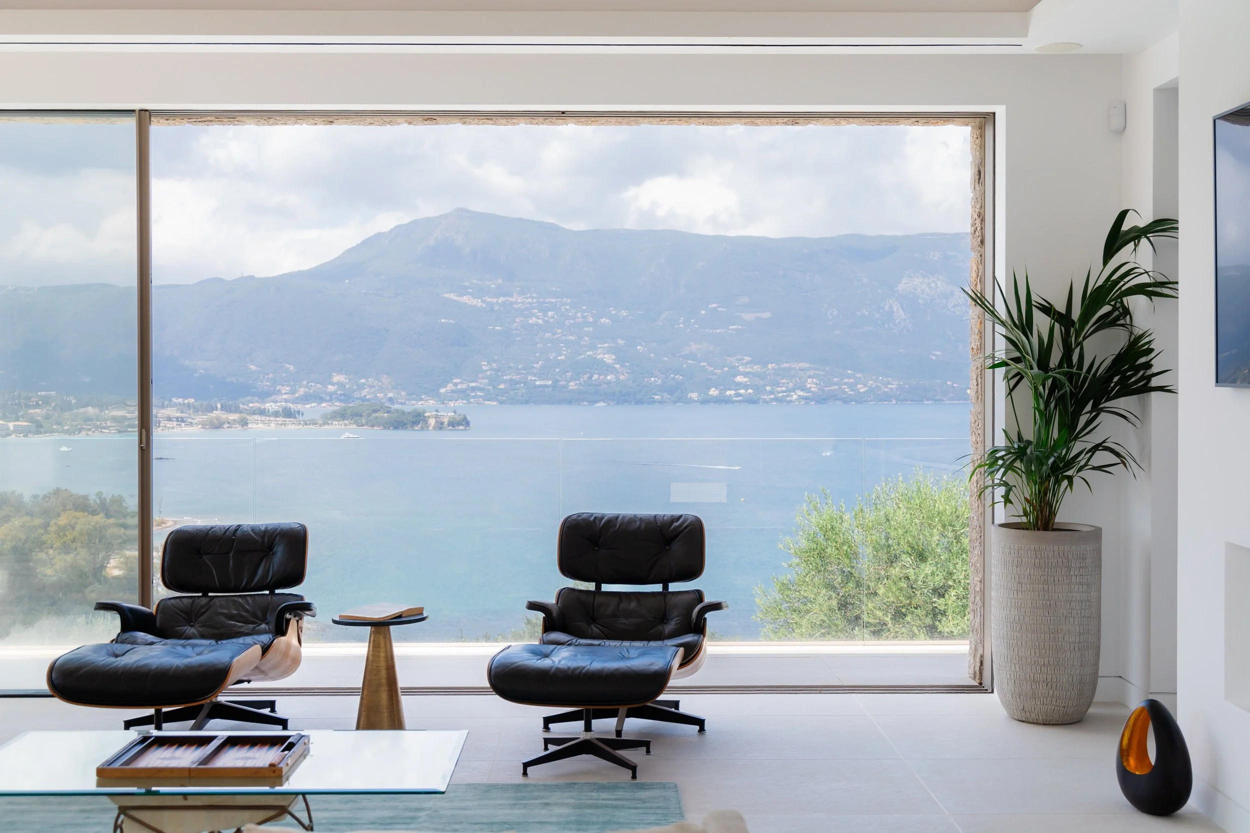 Modern living room with a large floor-to-ceiling window overlooking a lake and mountains, featuring two black leather chairs, a small side table, a potted plant, a wall-mounted TV, and minimal decor.